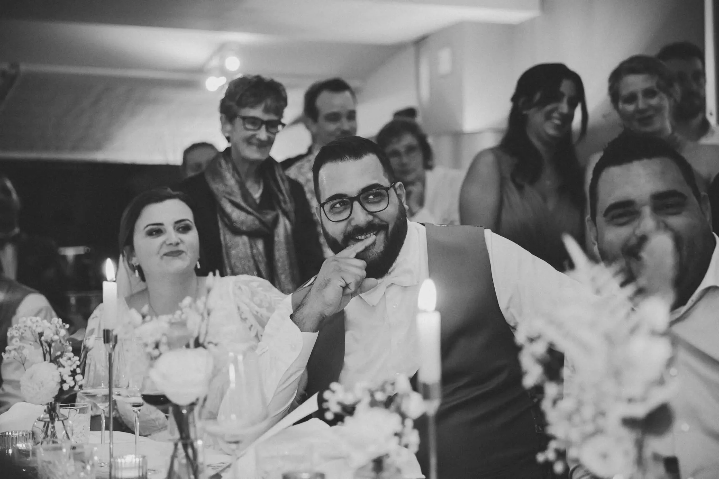 Menschen lachen und feiern bei einem festlichen Anlass, sitzend an einem festlich gedeckten Tisch mit Blumen und Kerzen.