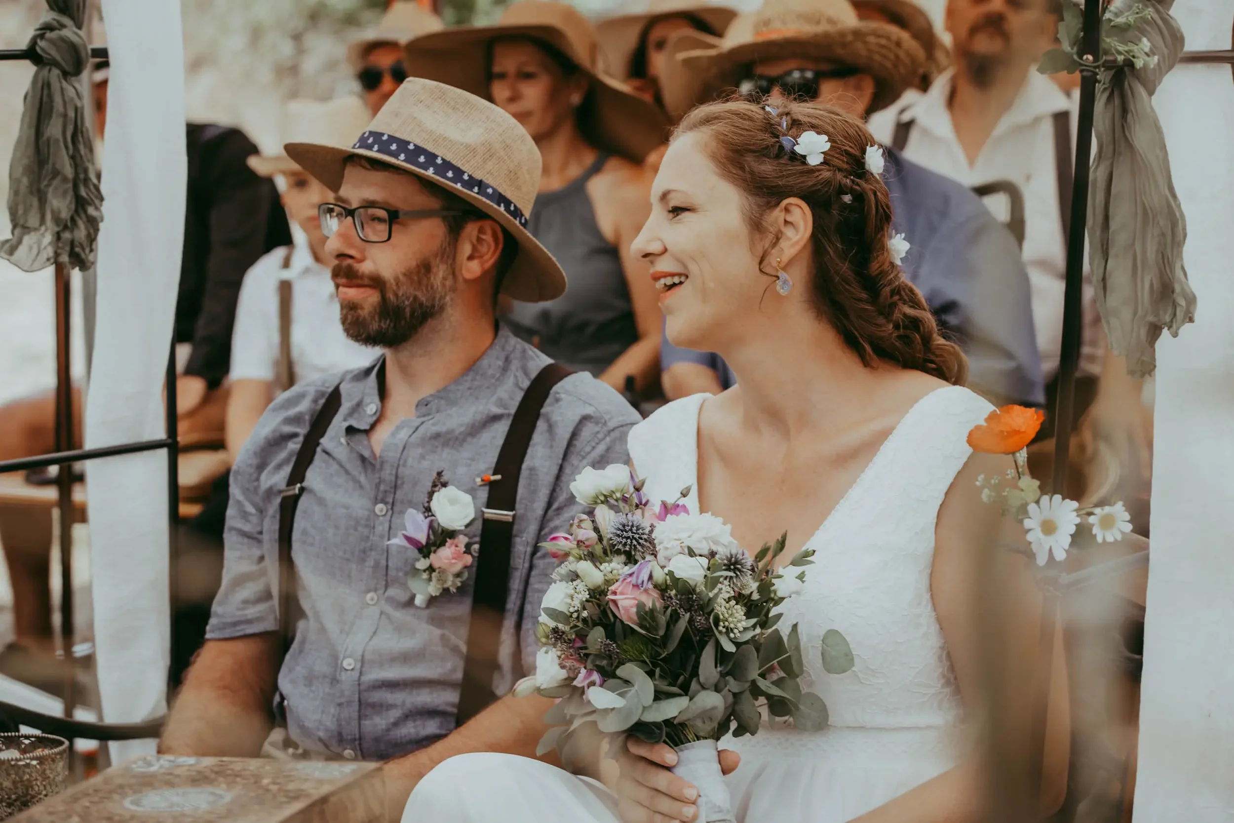 Ein lachendes Brautpaar bei einer Hochzeitsfeier, umgeben von Gästen, mit Brautstrauß und Blüten in den Haaren, im Freien bei Sonnenschein.