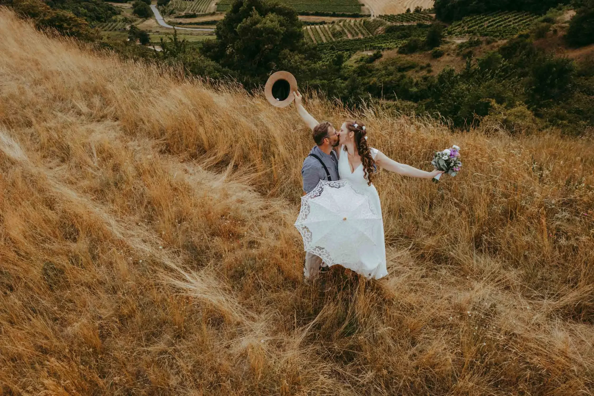 Ein Brautpaar steht auf einem Hügel mit goldenen Gräsern, die den Hintergrund bilden, während sie sich küssen. Die Braut trägt ein weißes Kleid und hält einen weißen Regenschirm sowie einen Blumenstrauß, der Bräutigam trägt einen blauen Hemd und hat 