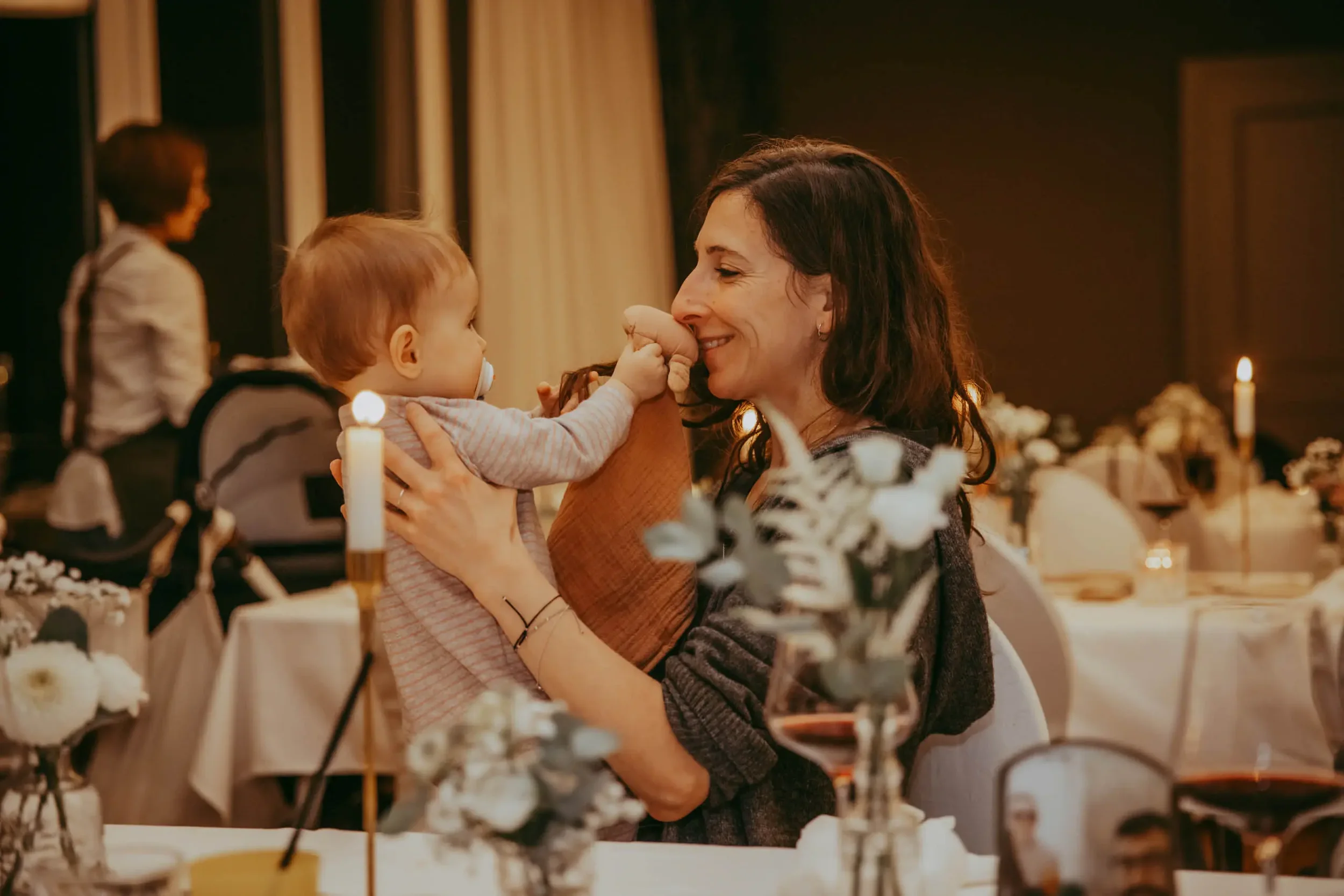 Eine Frau hält ein Baby in den Armen, beide lachen im Kerzenlicht bei einer Feier oder einem Abendessen, im Hintergrund weitere Personen und Kerzen.