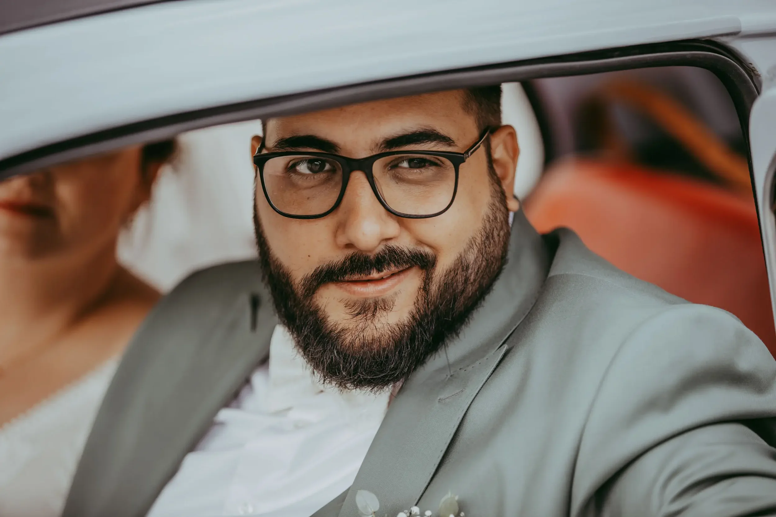 Nahaufnahme eines Mannes mit Bart und Brille, der aus einem Auto schaut und ein Lächeln zeigt.
