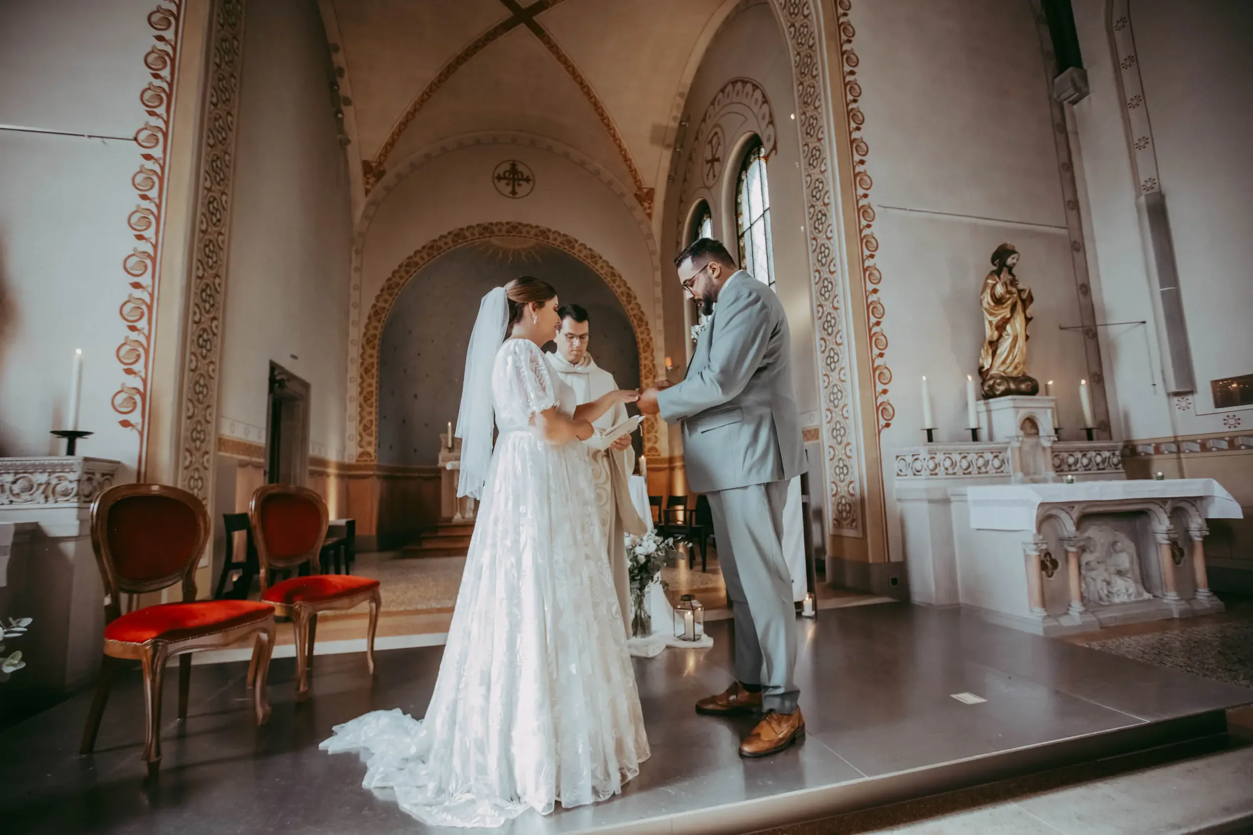 Ein Brautpaar bei ihrer standesamtlichen Trauung in einer Kirche, die sich küsst, während der Priester oder Trauzeugen im Hintergrund zuschaut.