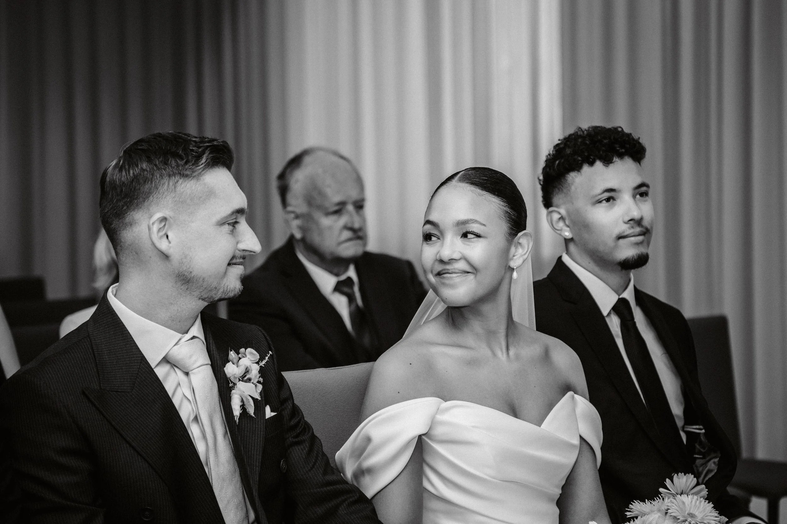 Schwarz-weiß Foto einer Hochzeit mit Braut und Bräutigam, sowie weitere Gäste, in einem Saal mit Vorhängen im Hintergrund.