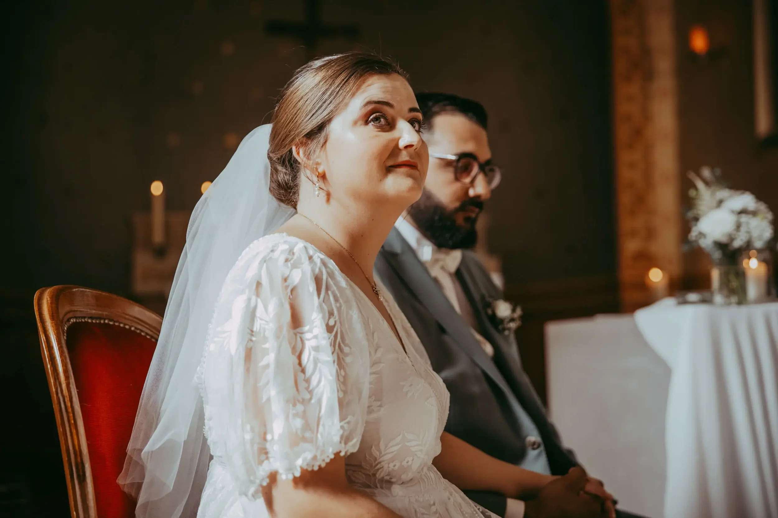 Braut und Bräutigam sitzen bei einer Hochzeitszeremonie in der Kirche, die Braut trägt ein weißes Kleid mit Spitze und einen Schleier, während der Bräutigam einen Anzug trägt.