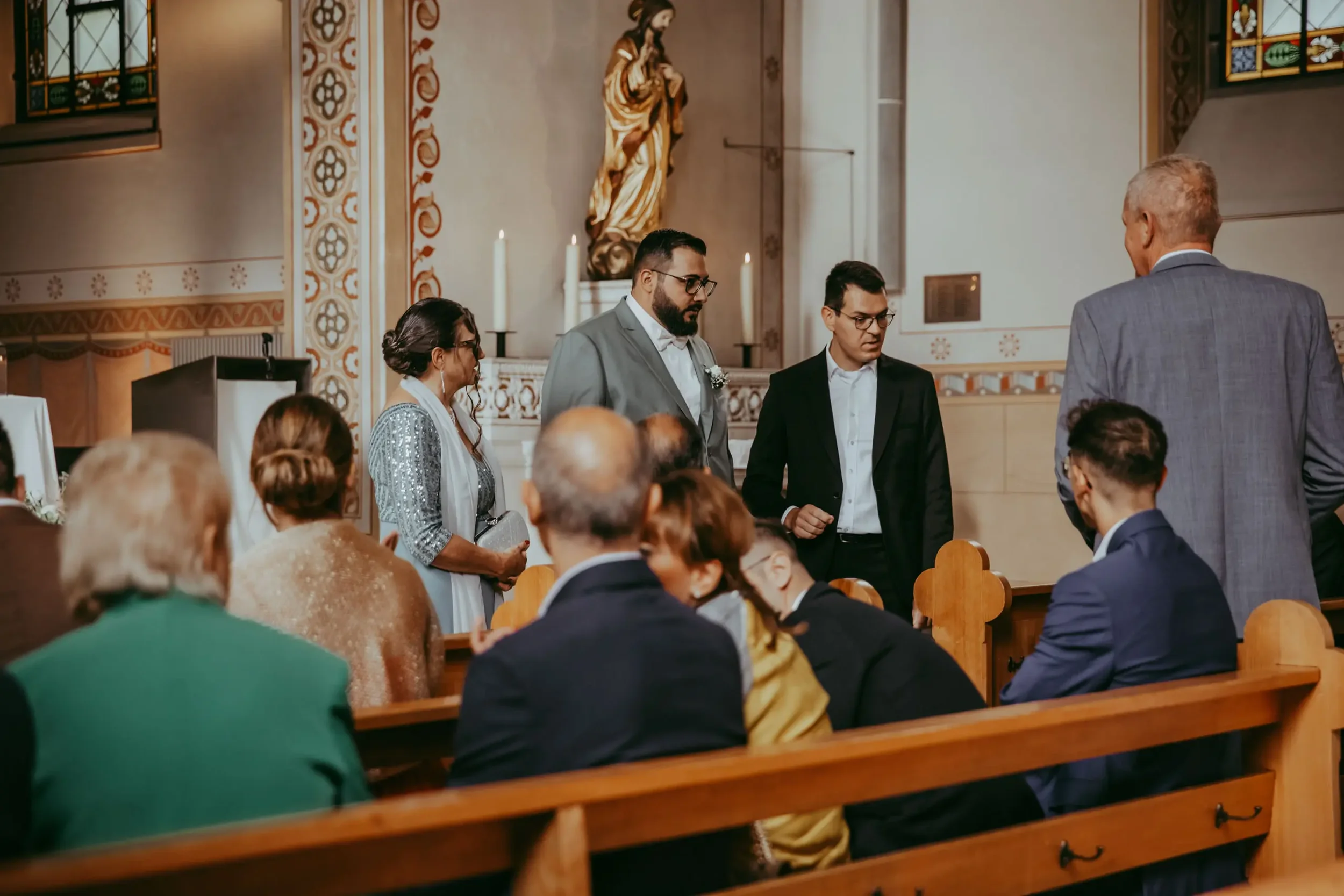 Menschen stehen in einer Kirche während einer Zeremonie, vermutlich eine Hochzeit, mit kunstvollen Wanddekorationen und religiösen Statuen im Hintergrund.