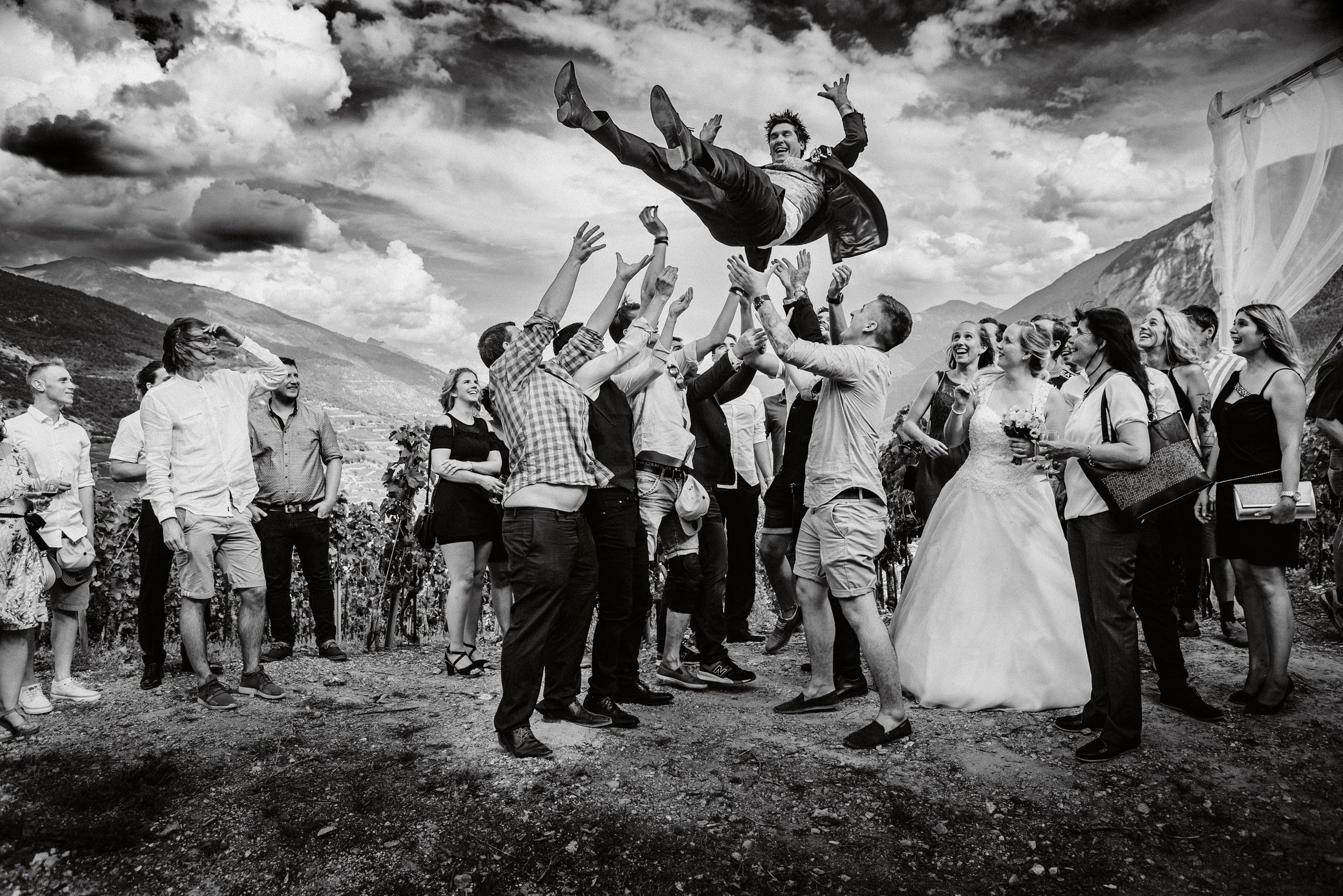 Hochzeit im Freien mit Hochzeitsgesellschaft, die Braut in Hochzeitskleid und Bräutigam feiern, während eine Person auf Händen getragen wird, umrahmt von Bergen und Wolken im Himmel.