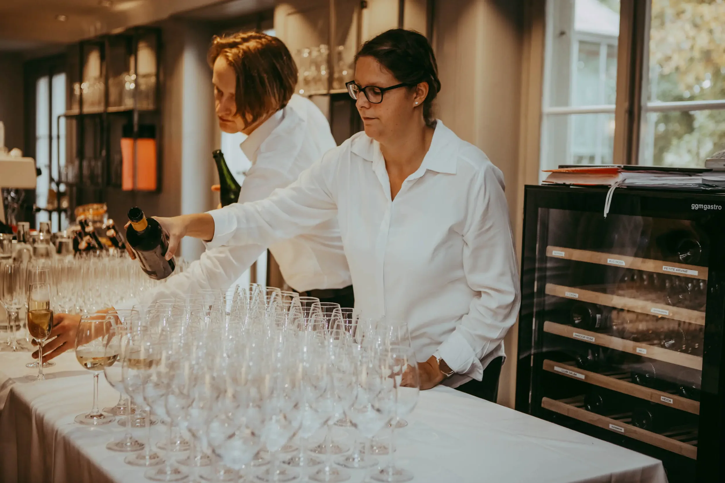 Zwei Frauen in weißen Hemden füllen Sektgläser bei einer Feier in einem Raum mit großen Fenstern und einem Weinregal im Hintergrund.