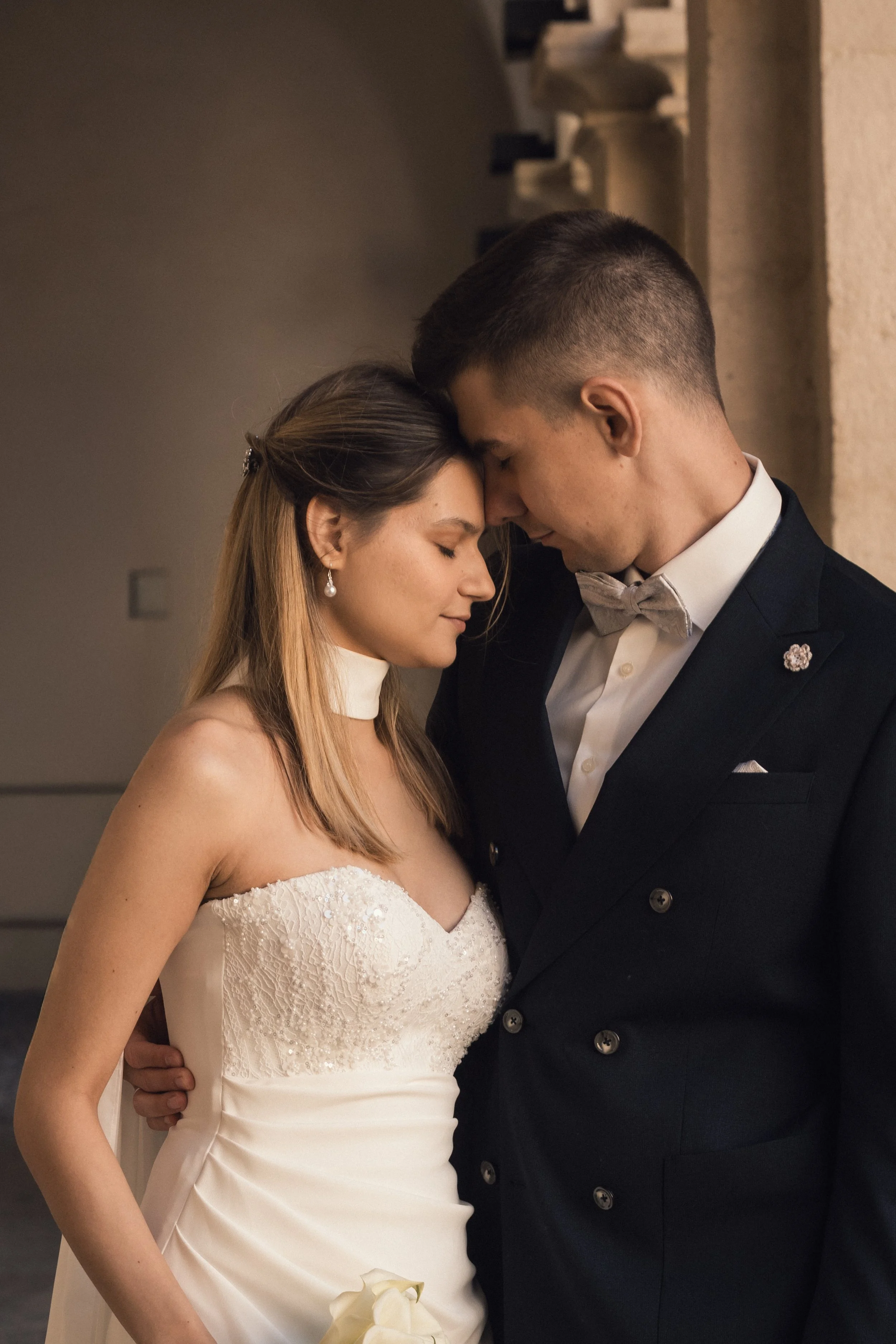 Braut und Bräutigam bei ihrer Hochzeit, stehen eng zusammen mit den Stirnen aneinander, im Hintergrund eine Steinmauer.