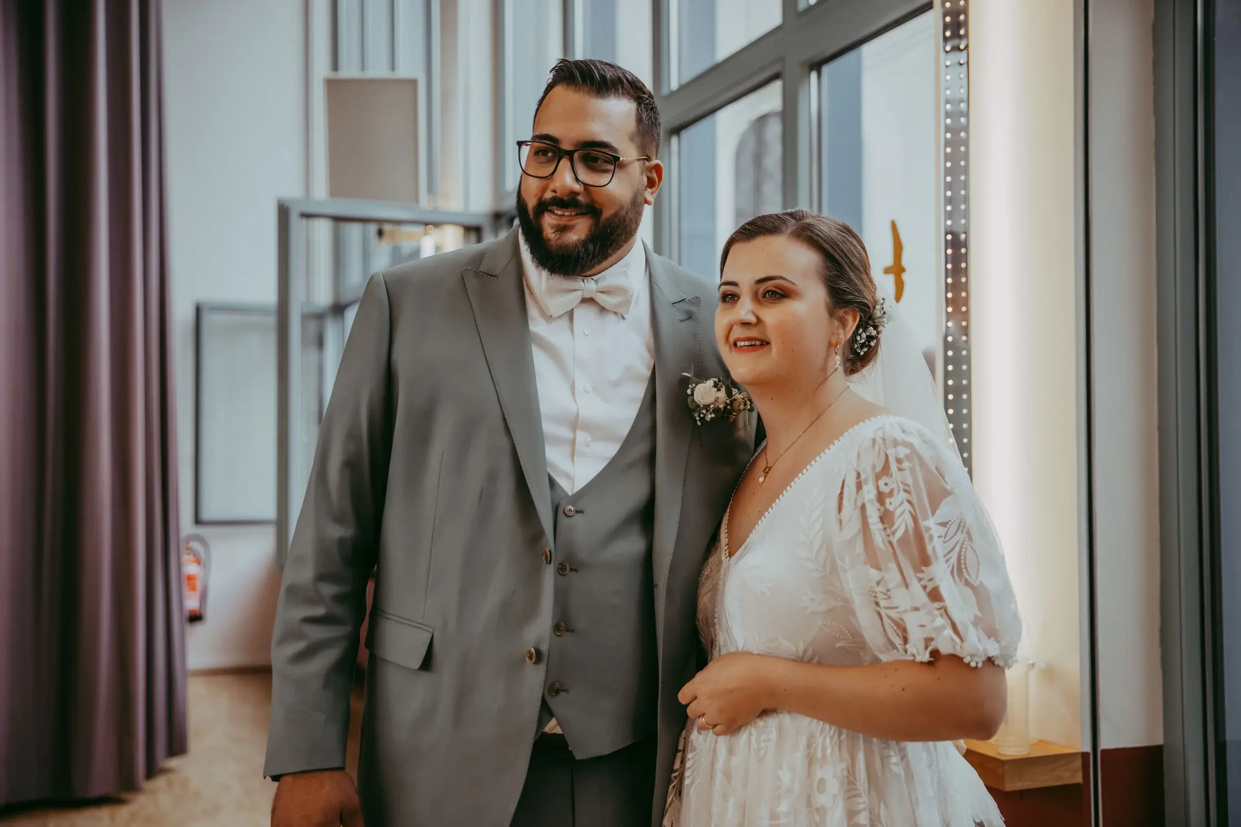 Ein Mann in einem grauen Anzug mit Brille und einer Frau in einem weißen Brautkleid stehen in einem Aufzug und lächeln.