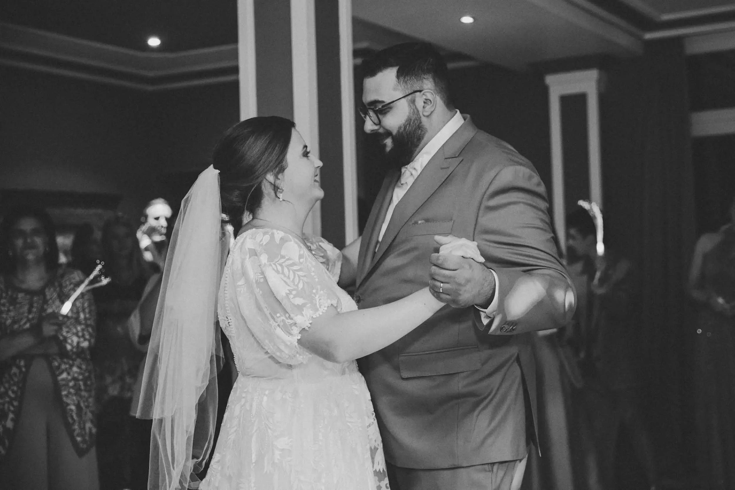 Ein Brautpaar tanzt bei einer Hochzeit. Sie lächeln einander an, während sie sich halten. Im Hintergrund sind Gäste, die zuschauen.