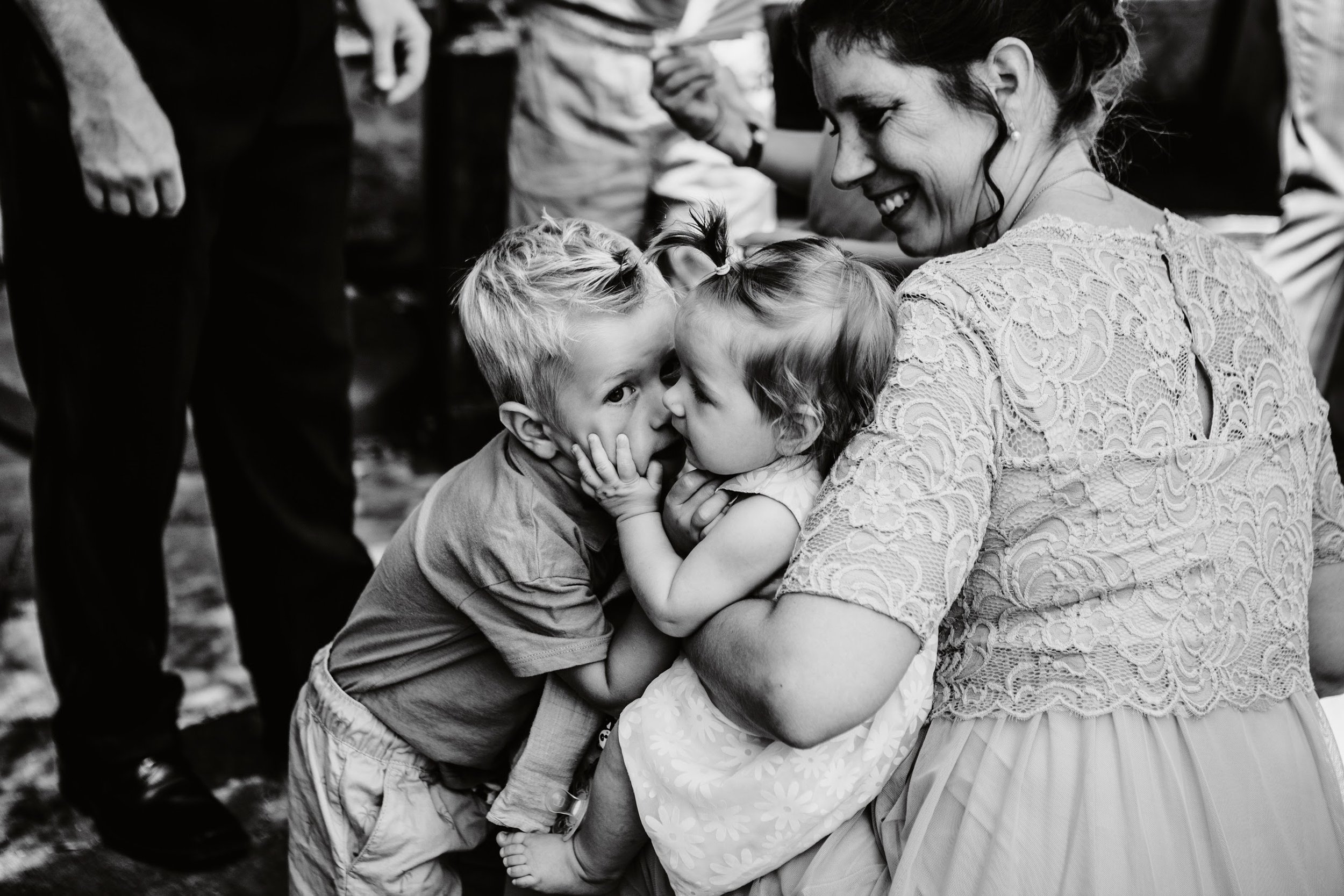 Eine Frau hält zwei kleine Kinder im Arm, küssend und lächelnd, während eine Gruppe von Menschen im Hintergrund steht.