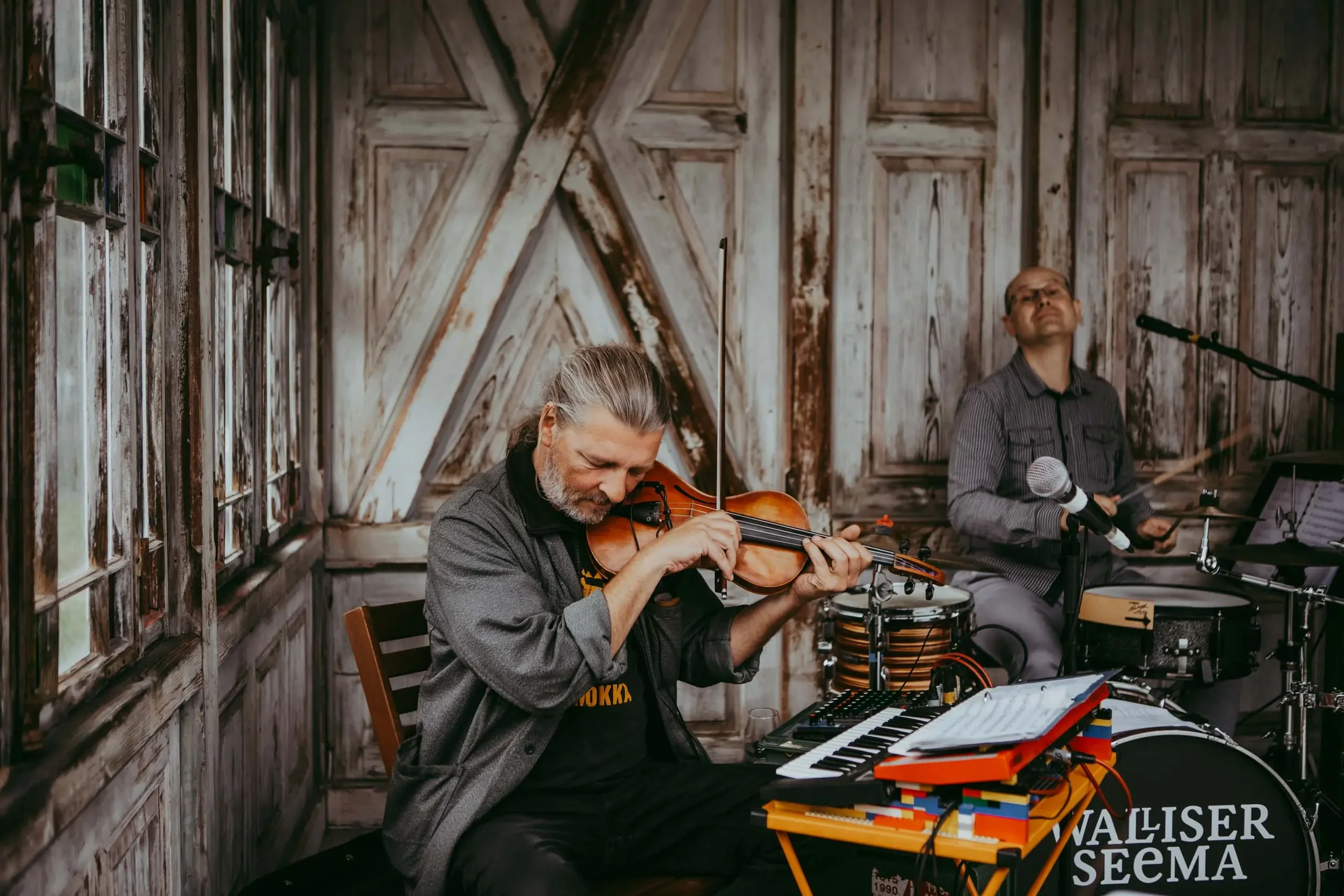Zwei Musiker spielen in einem rustikalen Holzraum auf verschiedenen Instrumenten, darunter Geige, Schlagzeug und Keyboard.