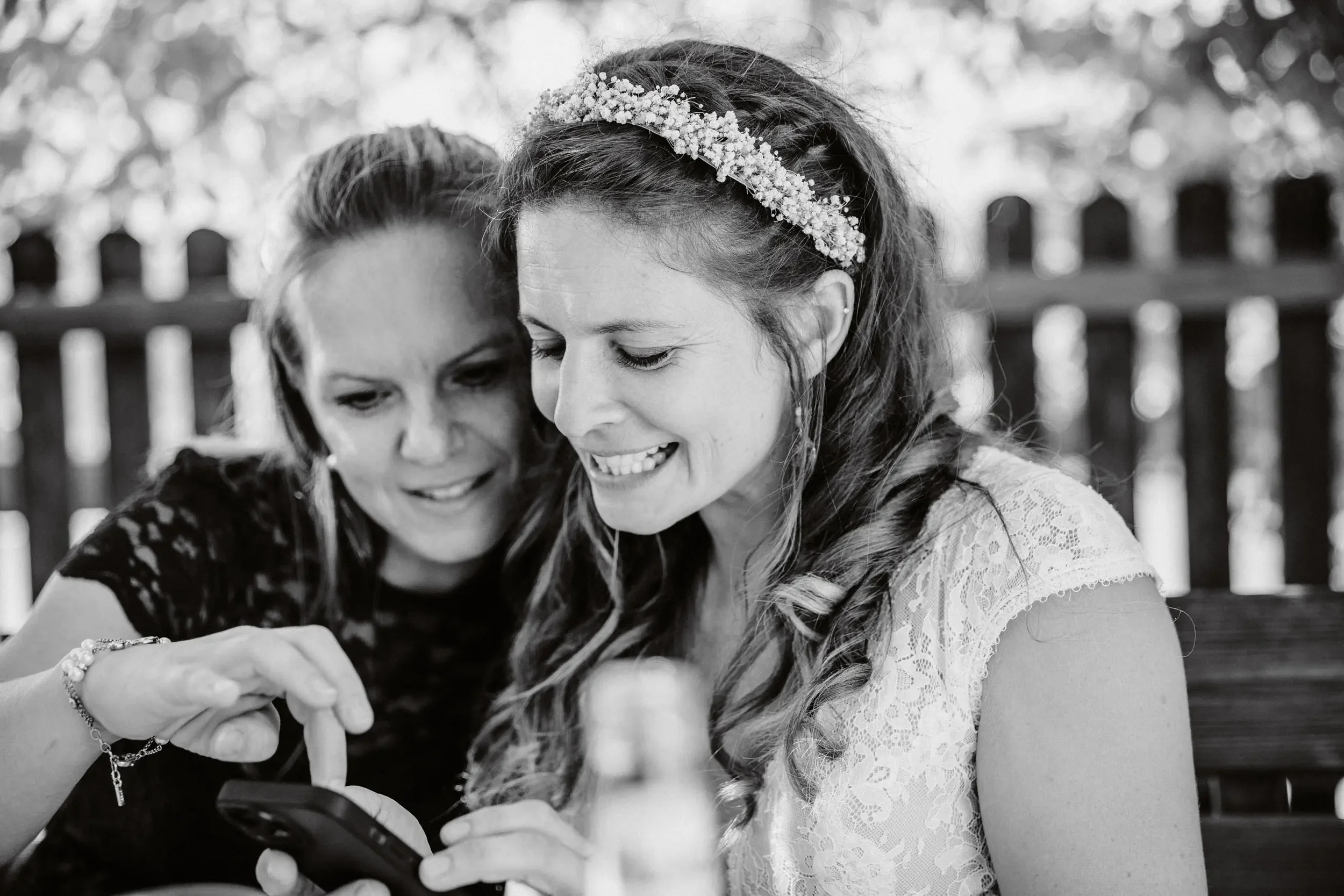 Zwei Frauen, eine älter, die andere jünger, schauen zusammen auf ein Smartphone. Die jüngere Frau trägt eine Blume Kopfhaube, wahrscheinlich für eine Hochzeit.