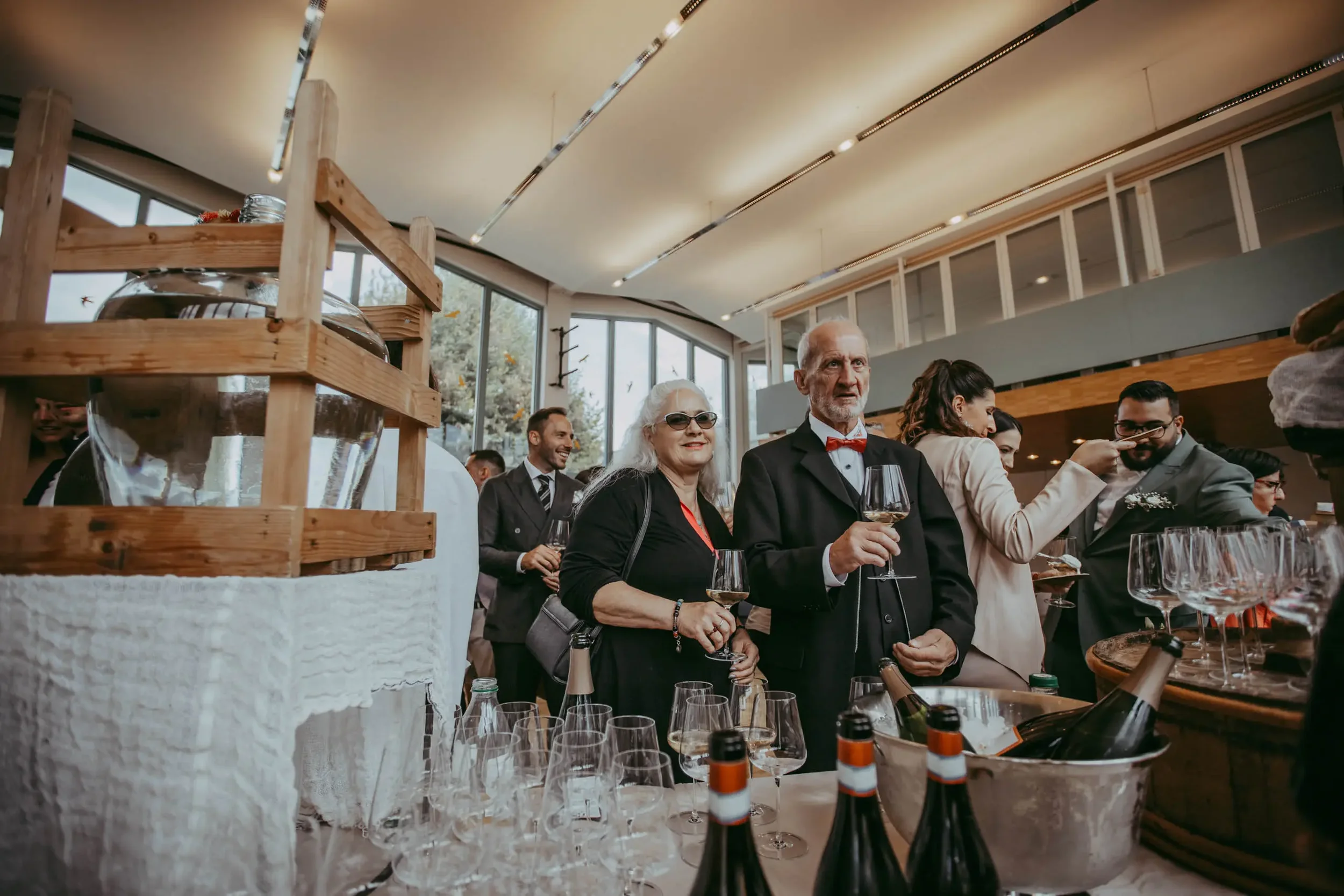 Ältere Frau mit weißen Haaren und Sonnenbrille, jüngerer Mann mit weißen Haaren und roter Fliege, beide halten Weingläser, auf einem festlich gedeckten Tisch bei einer Feier im Restaurant.