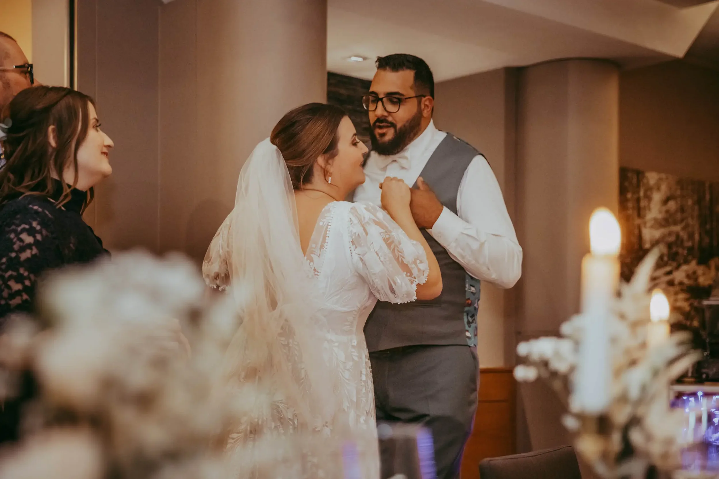 Ein Brautpaar bei einer Hochzeit spricht an einem festlichen Ort, während eine Frau lächelnd zusieht.