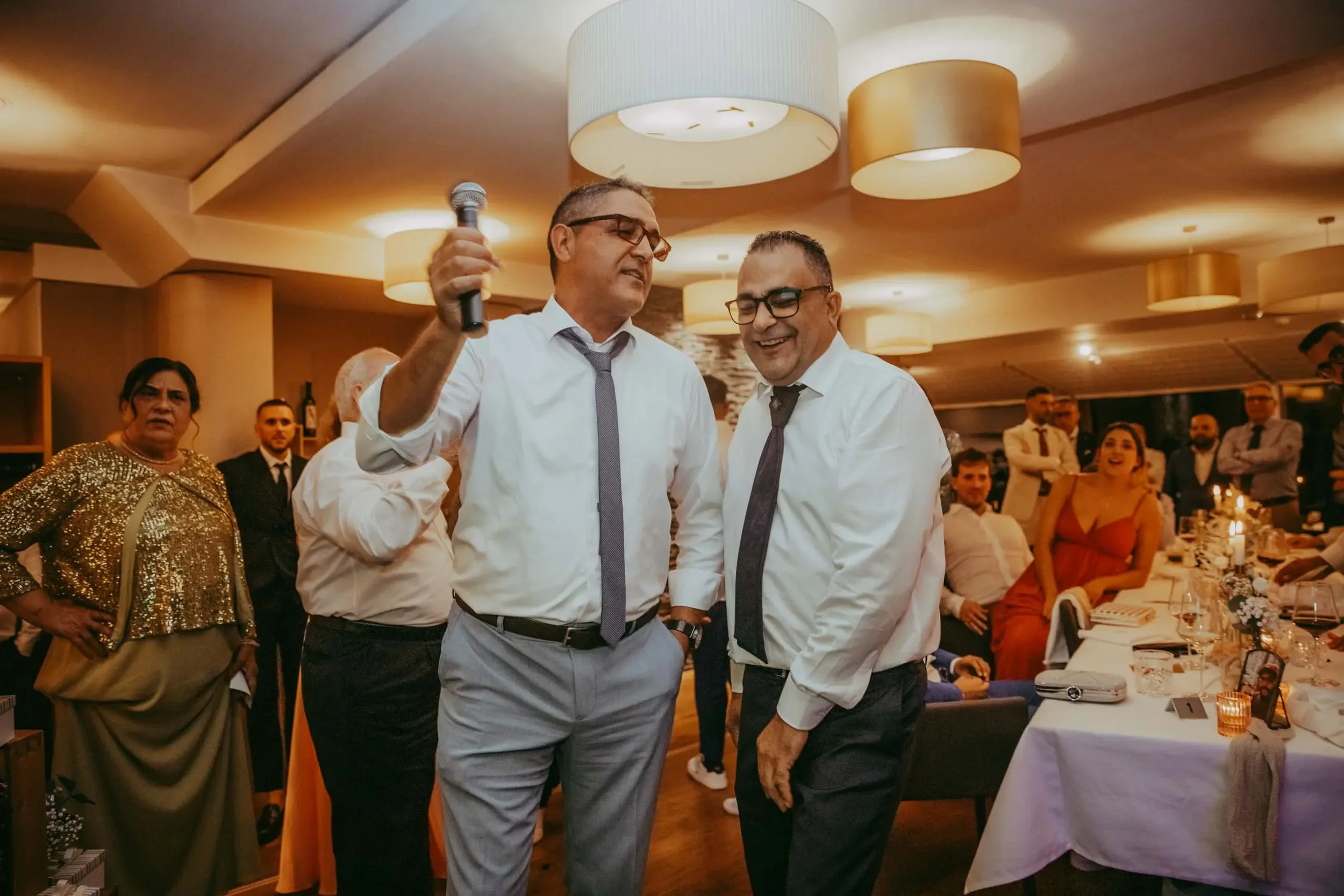 Zwei Männer in formeller Kleidung unterhalten sich auf einer Feier in einem eleganten Raum, während andere Gäste im Hintergrund sitzen.