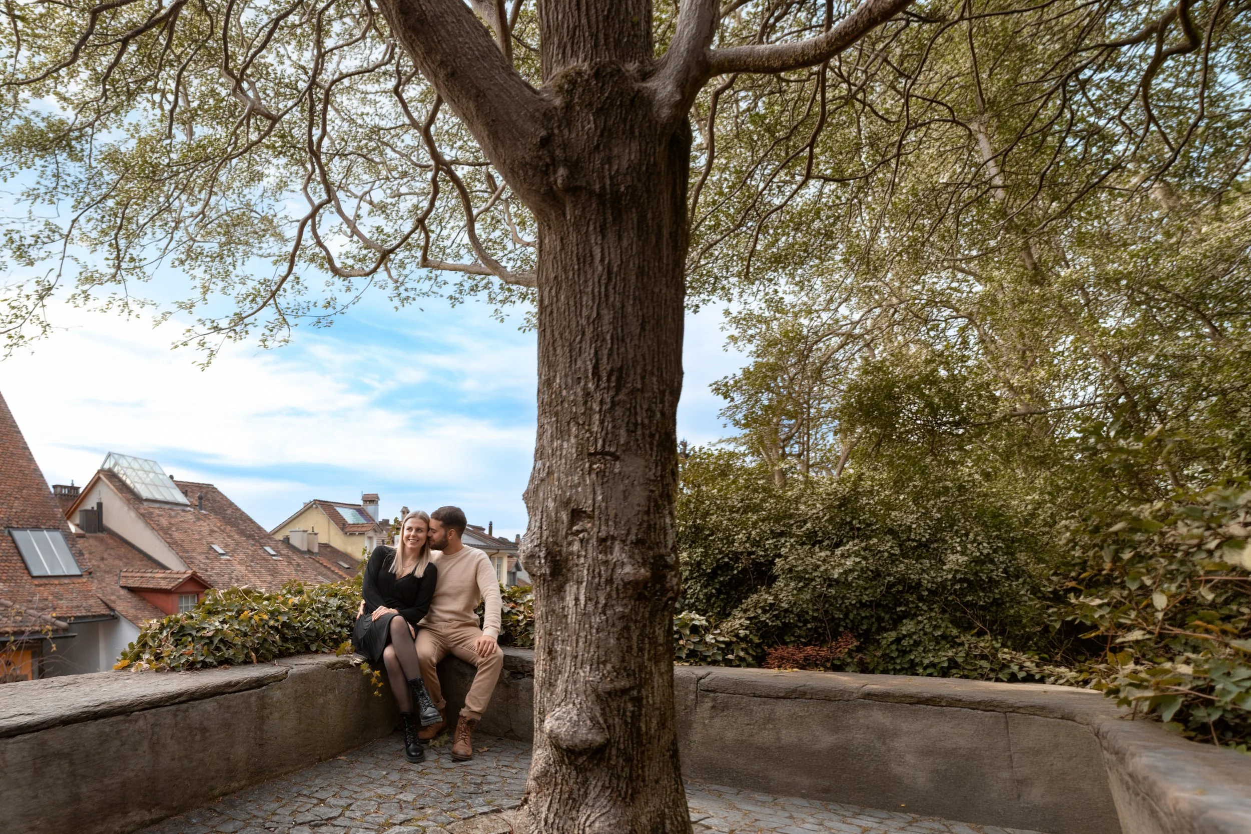 Paar sitzt auf einer Mauer vor einem großen Baum in einem Stadtpark, im Hintergrund Dächer von Häusern.
