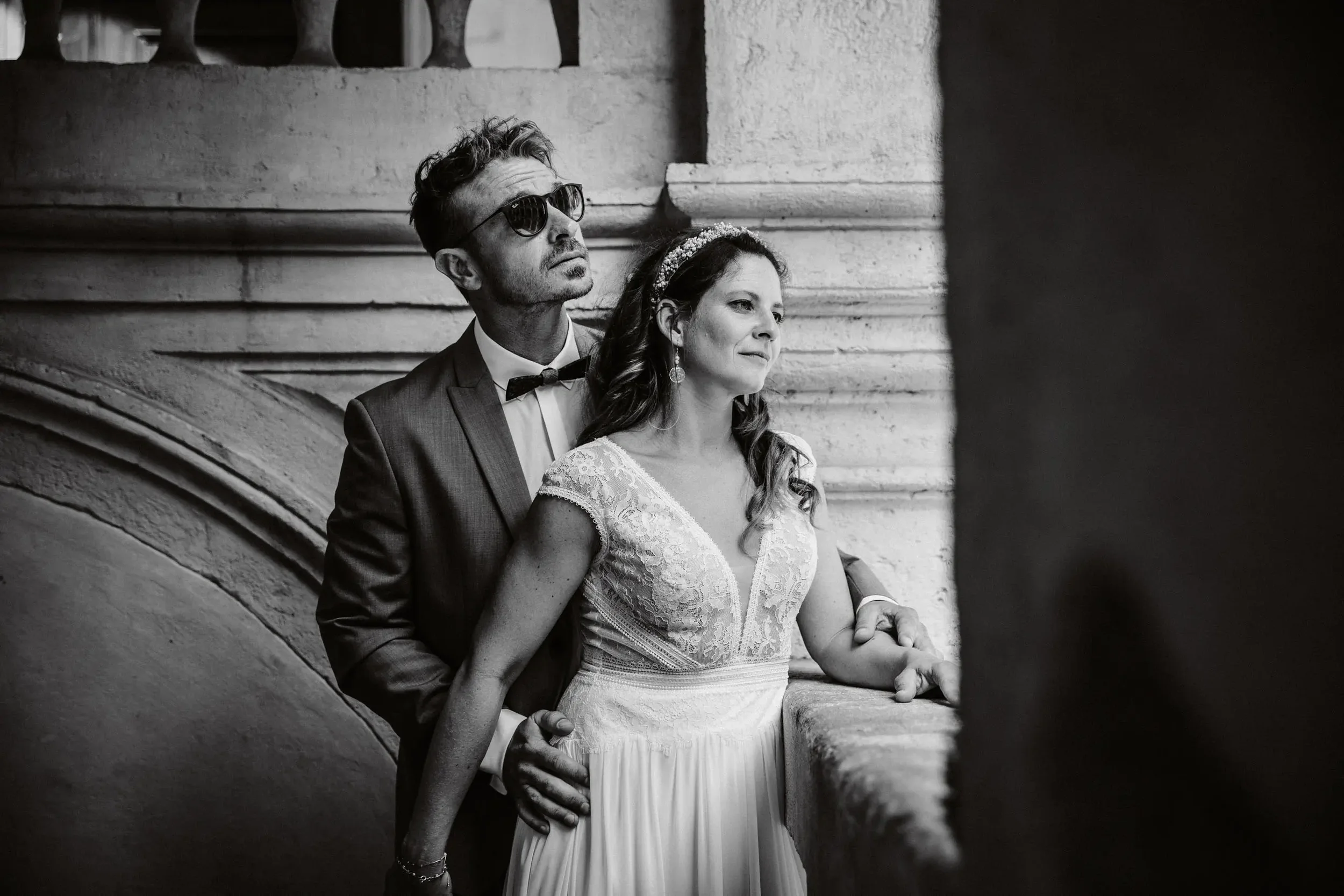 Schwarz-weiß-Foto eines Bräutigams in Sonnenbrille und Anzug, der hinter einer Braut in einem weißen Brautkleid steht, beide lehnen an einer Steinmauer, bei einer Hochzeitsaufnahme.