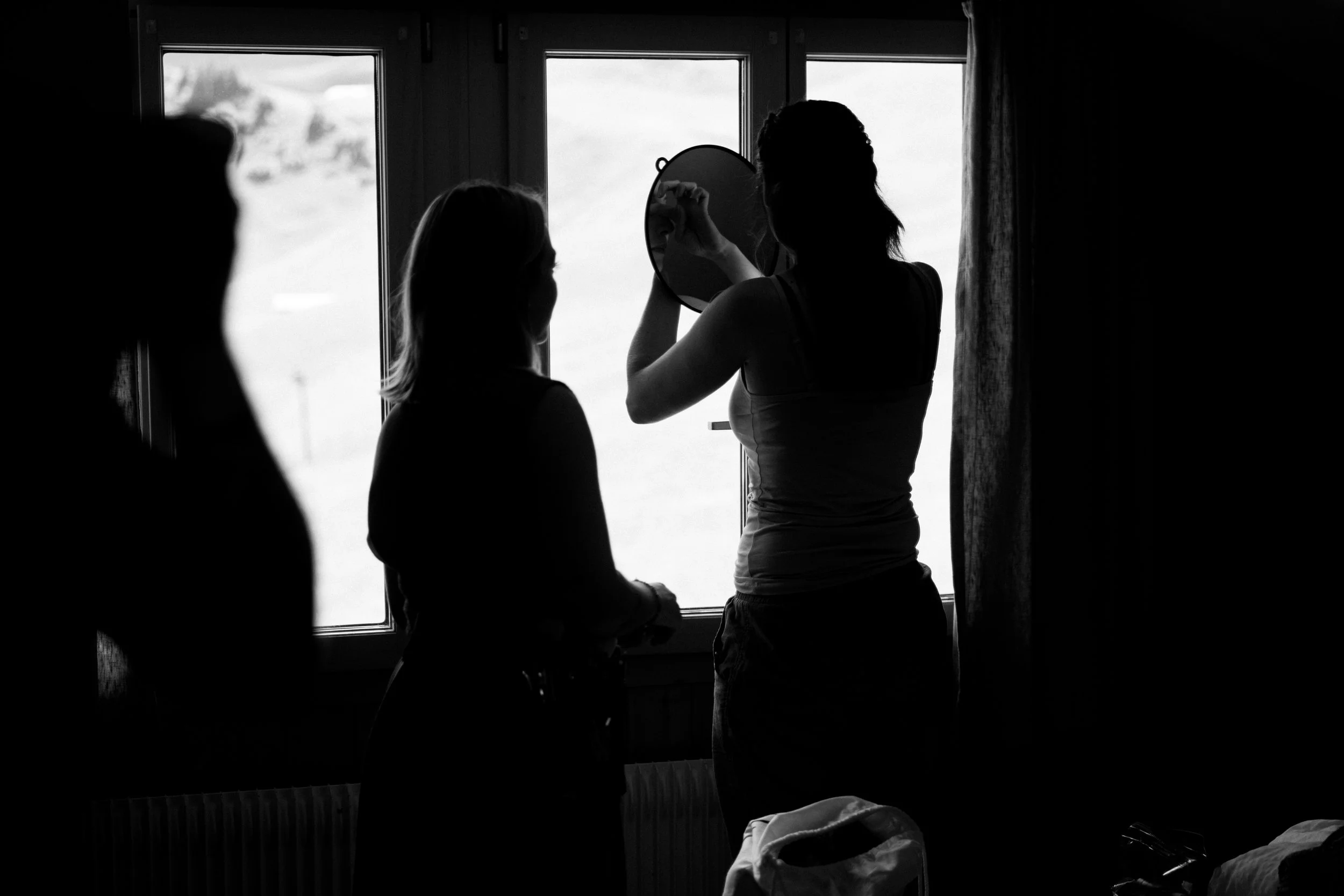 Drei Frauen vor einem Fenster, eine betrachtet sich im Spiegel, in Schwarz-Weiß-Fotografie.