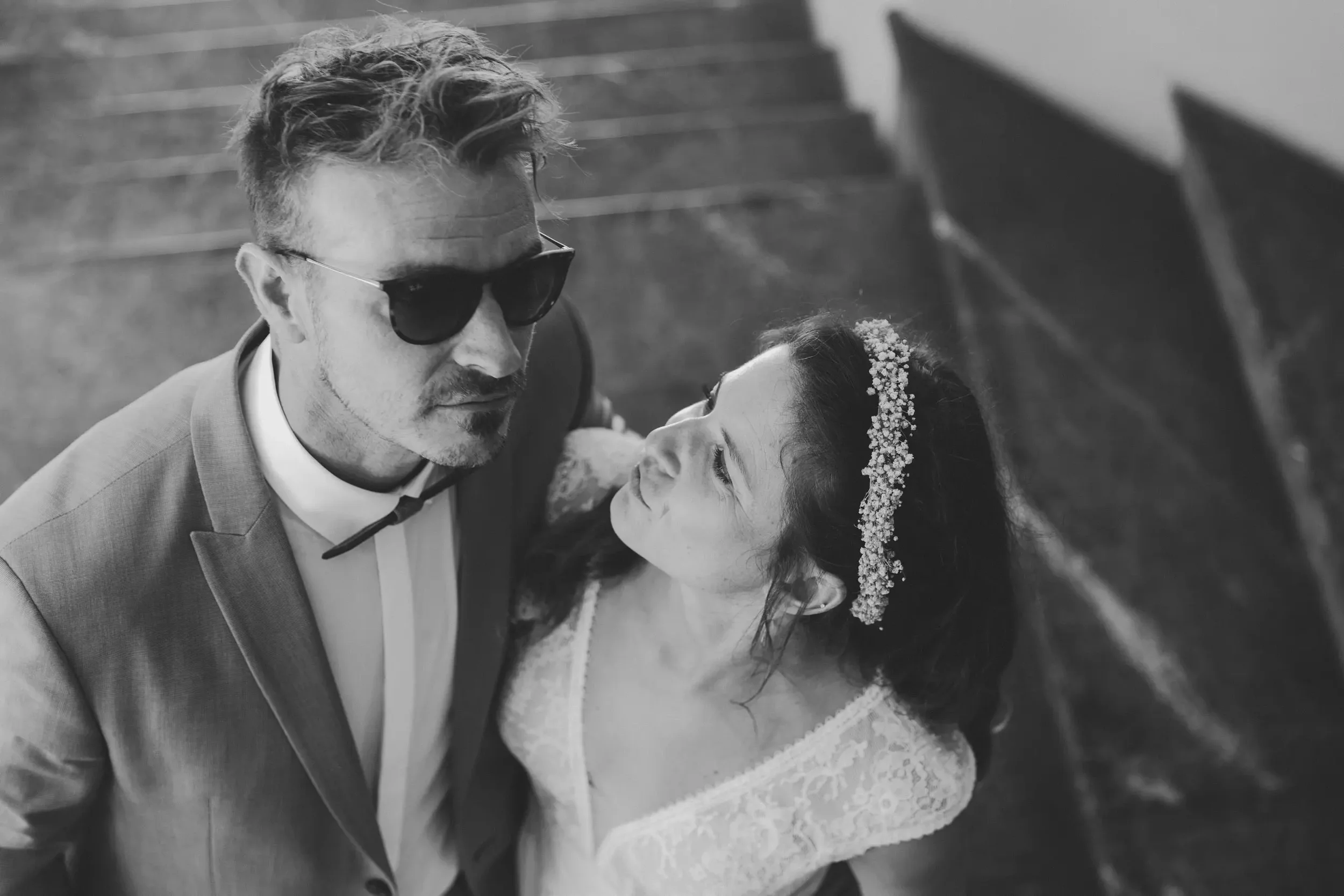 Ein Mann in Anzug und Sonnenbrille schaut eine Frau mit Schleier und Blumen im Haar an, beide stehen im Freien auf einer Treppe.