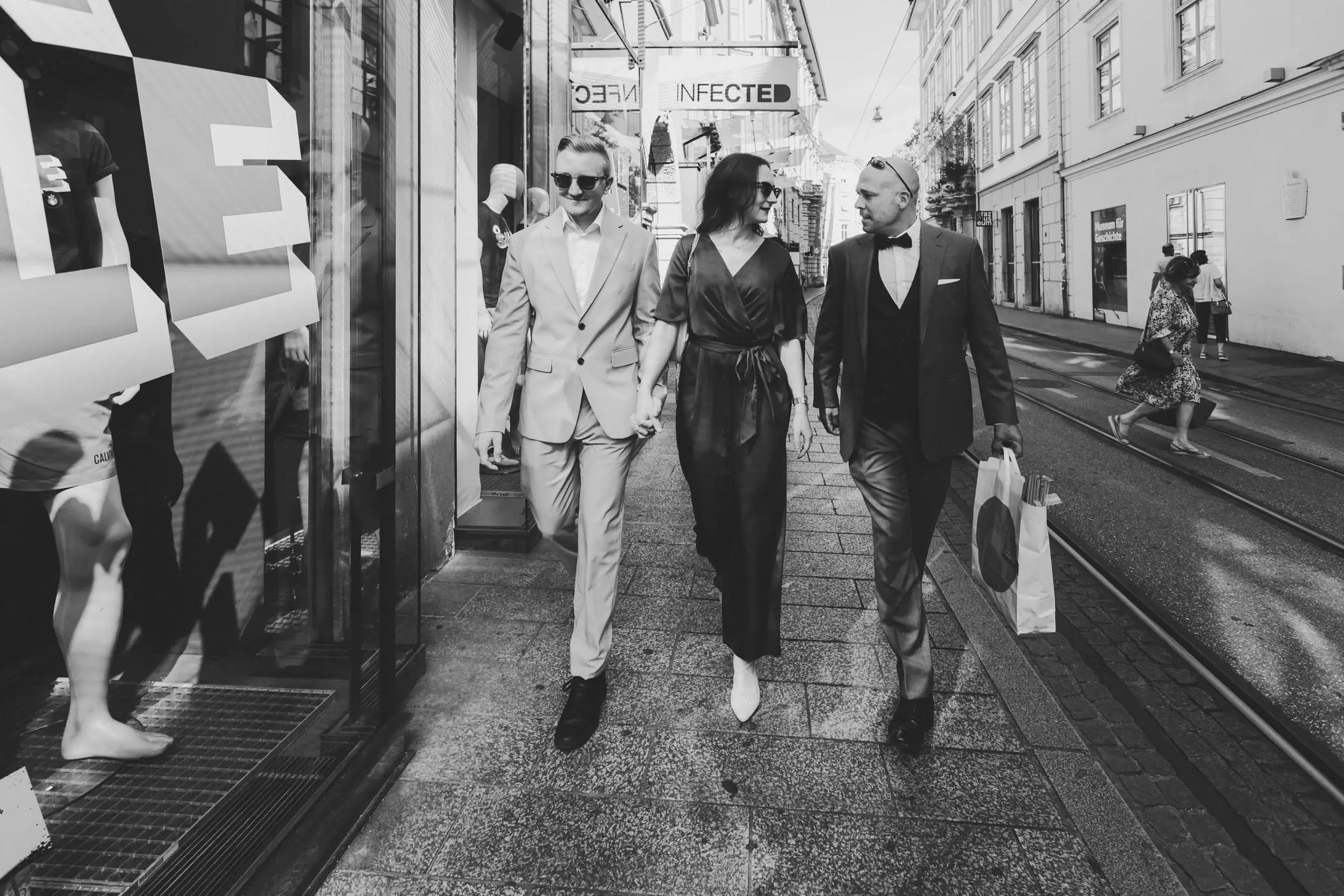 Drei elegante Menschen, zwei Männer und eine Frau, gehen Hand in Hand auf einer Stadtstraße, während im Hintergrund andere Passanten vorbeigehen. Es ist ein sonniger Tag, und sie tragen formelle Kleidung, die auf eine besondere Anlass oder einen stil