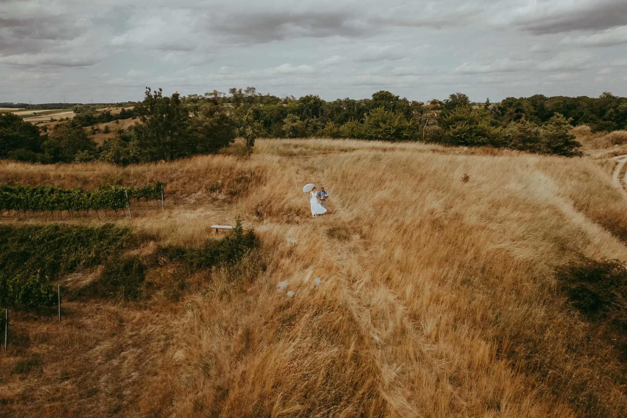 Eine Frau in einem weißen Kleid mit Regenschirm, die mit einem Mann in einem Karo-Hemd auf einem Feldweg in einer ländlichen Umgebung spazieren geht, umgeben von hohem, goldenem Gras, mit einem bewölkten Himmel im Hintergrund.