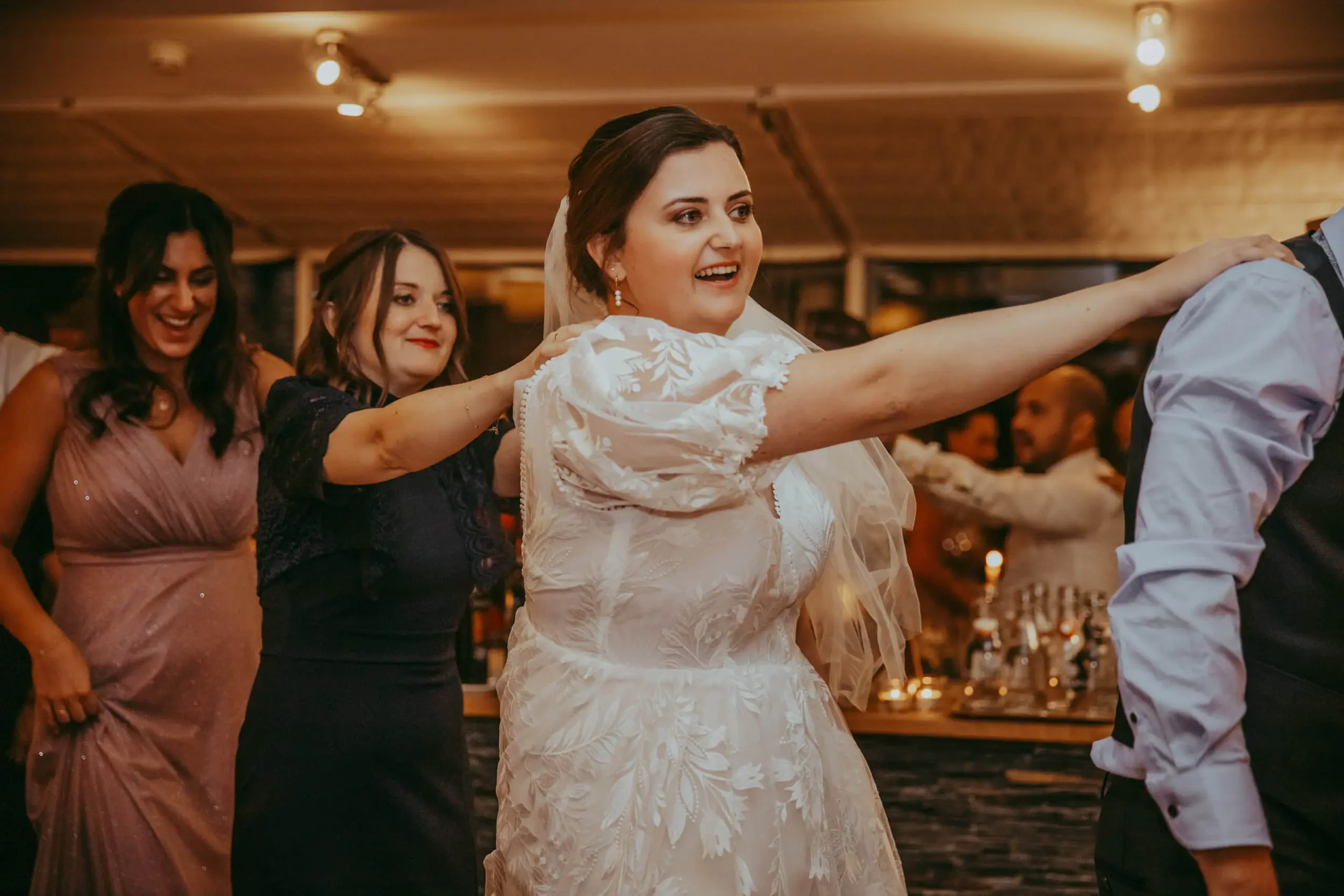 Hochzeitspaar tanzt, umgeben von Frauen und Gästen in einem festlichen Raum.