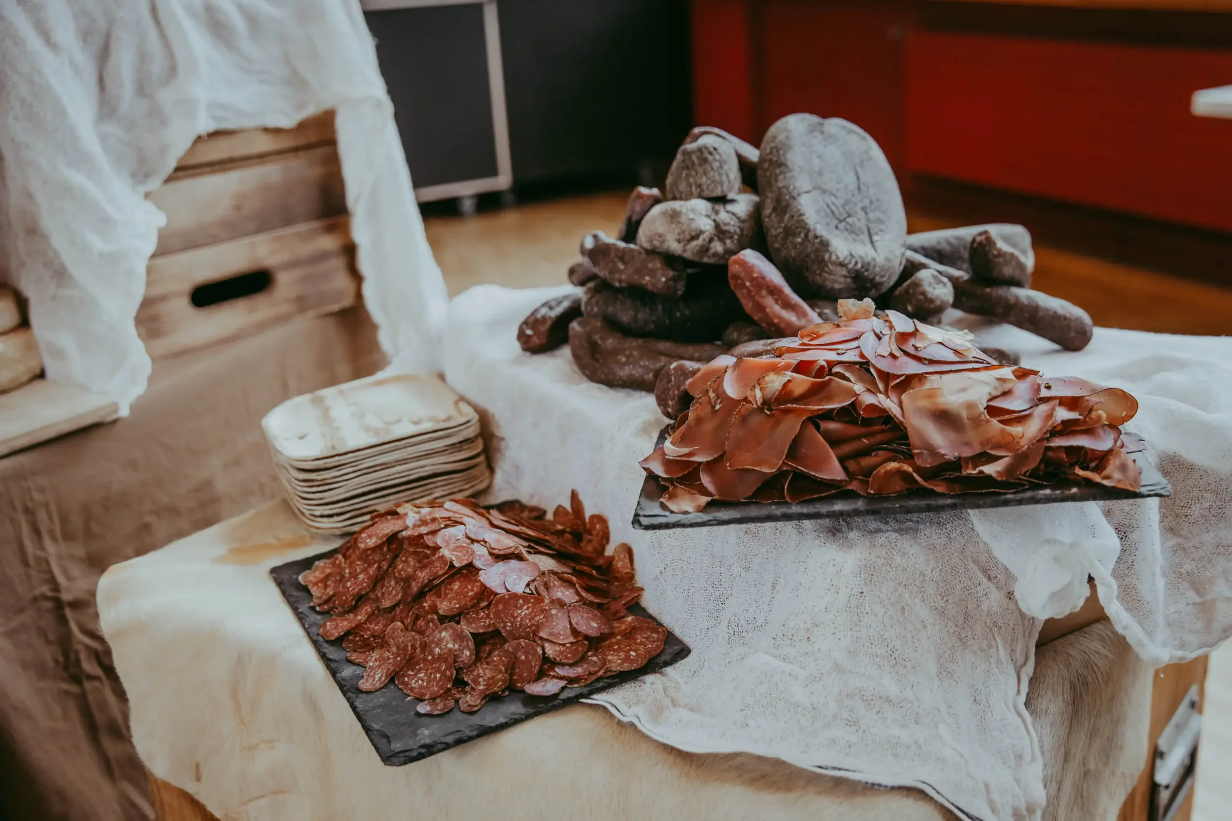 Auf einem Tisch sind verschiedene Wurstwaren und Schinken auf schwarzen Tellern angerichtet, daneben liegen Papiertücher.