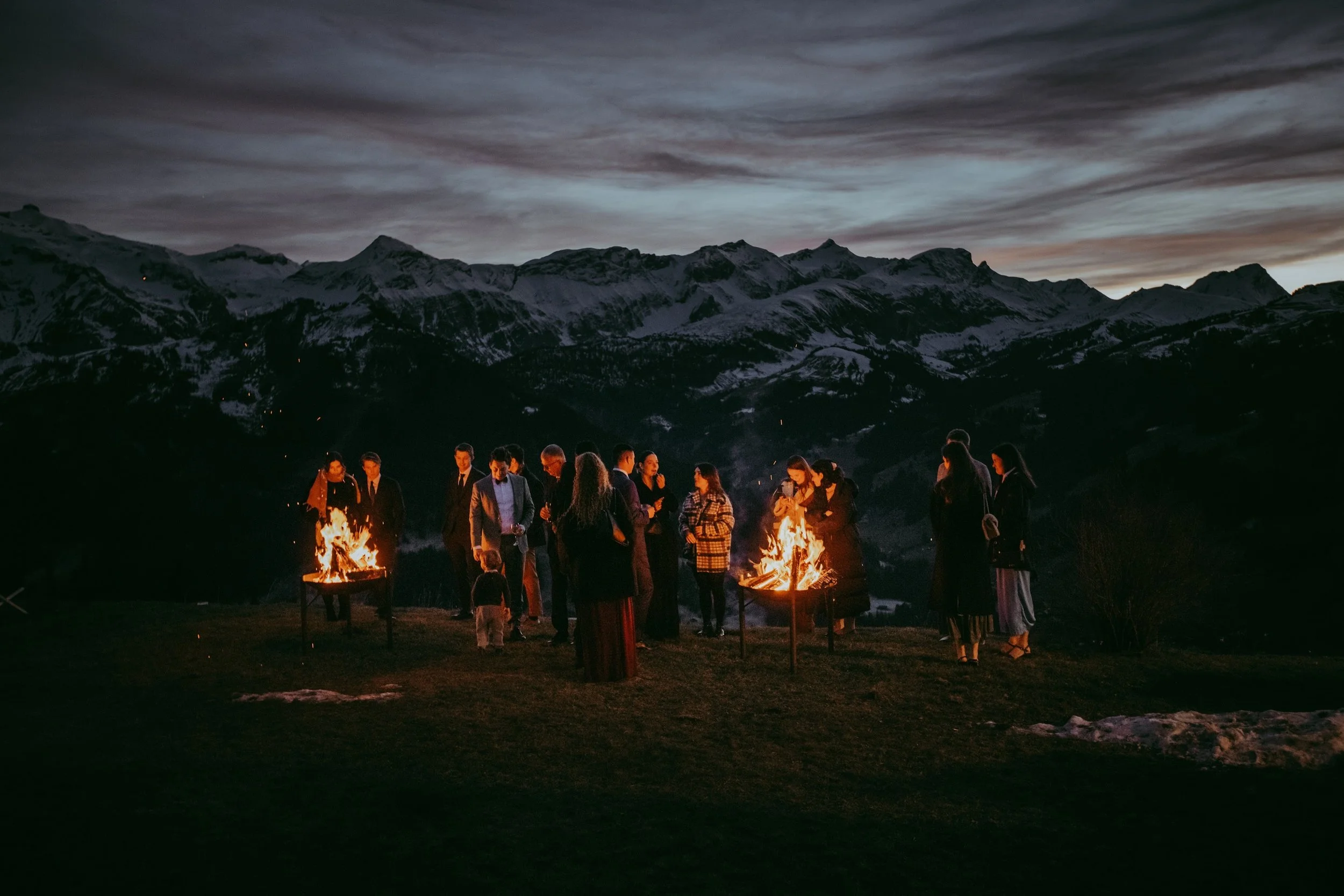 Menschen versammeln sich beim Lagerfeuer in einer nächtlichen Landschaft mit schneebedeckten Bergen im Hintergrund, bewölktem Himmel und vielen Wolken.