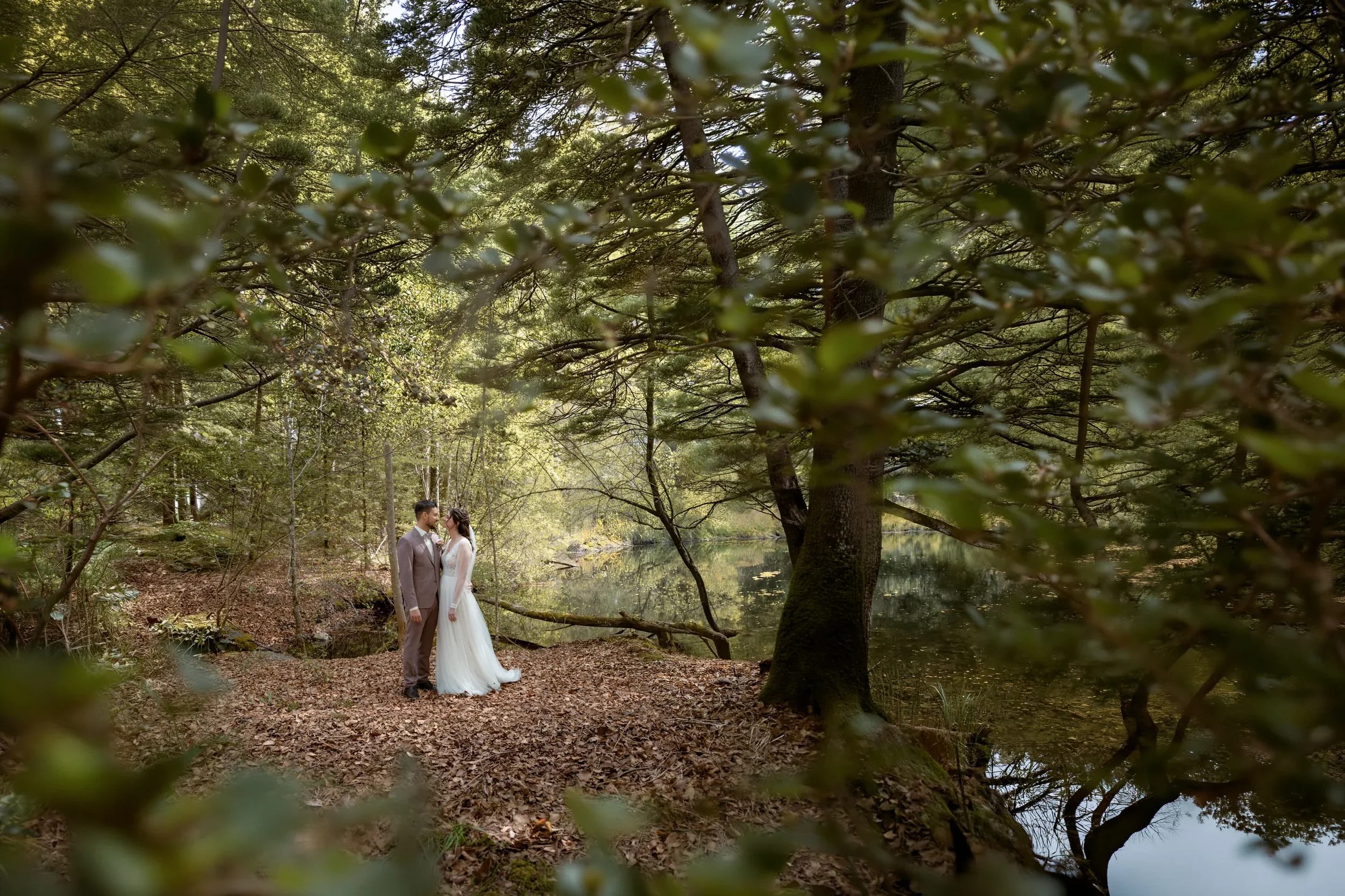 Ein Brautpaar steht in einem Wald am Fluss und schaut sich an, umgeben von grünen Bäumen und Sonnenlicht, das durch die Blätter fällt.