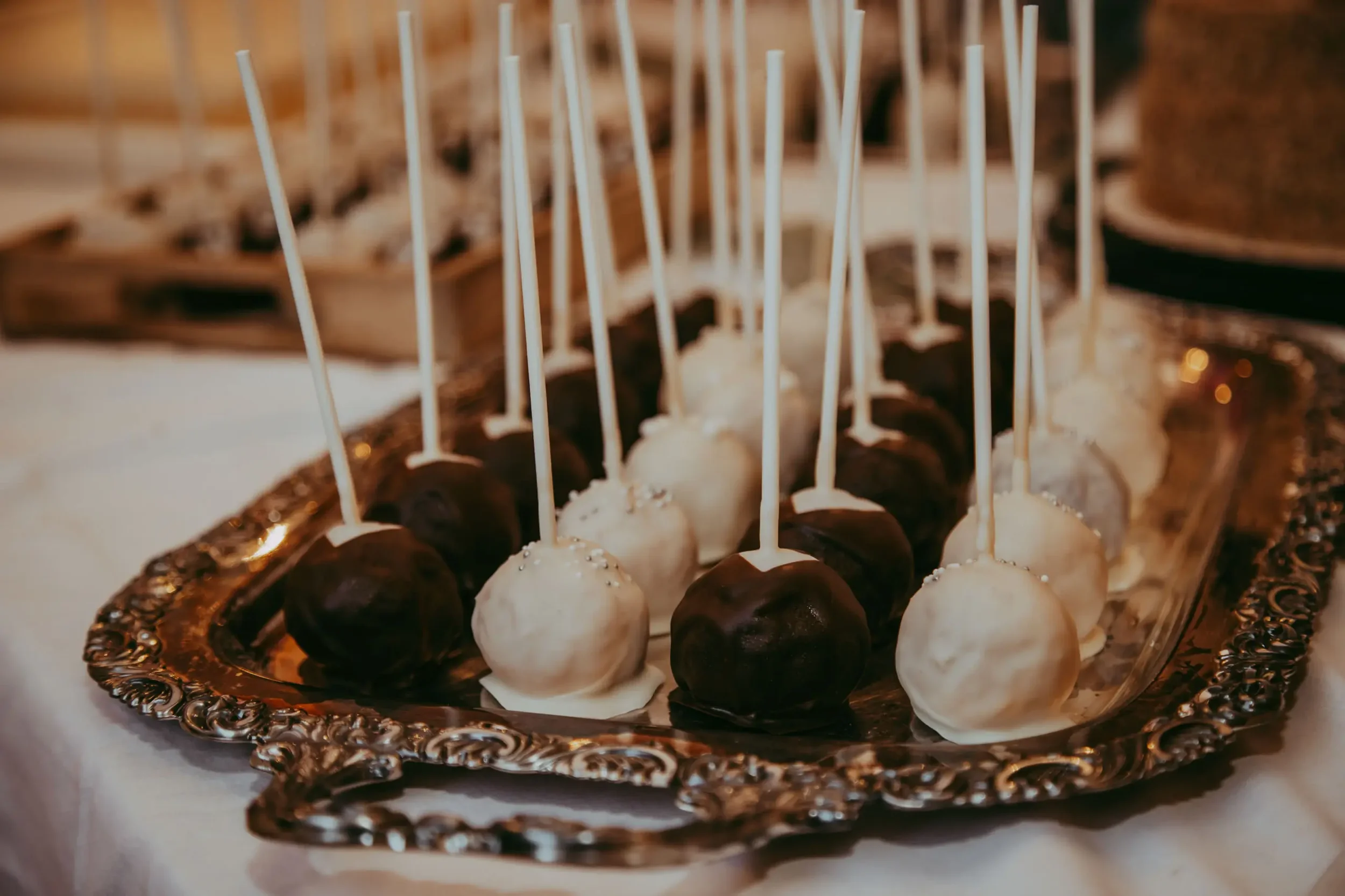 Dessertplatte mit runden Schokoladen- und Vanille-Cheeseballs, die auf Holzstäbchen gesteckt sind, auf einer dekorativen silbernen Platte mit Schnörkeln.