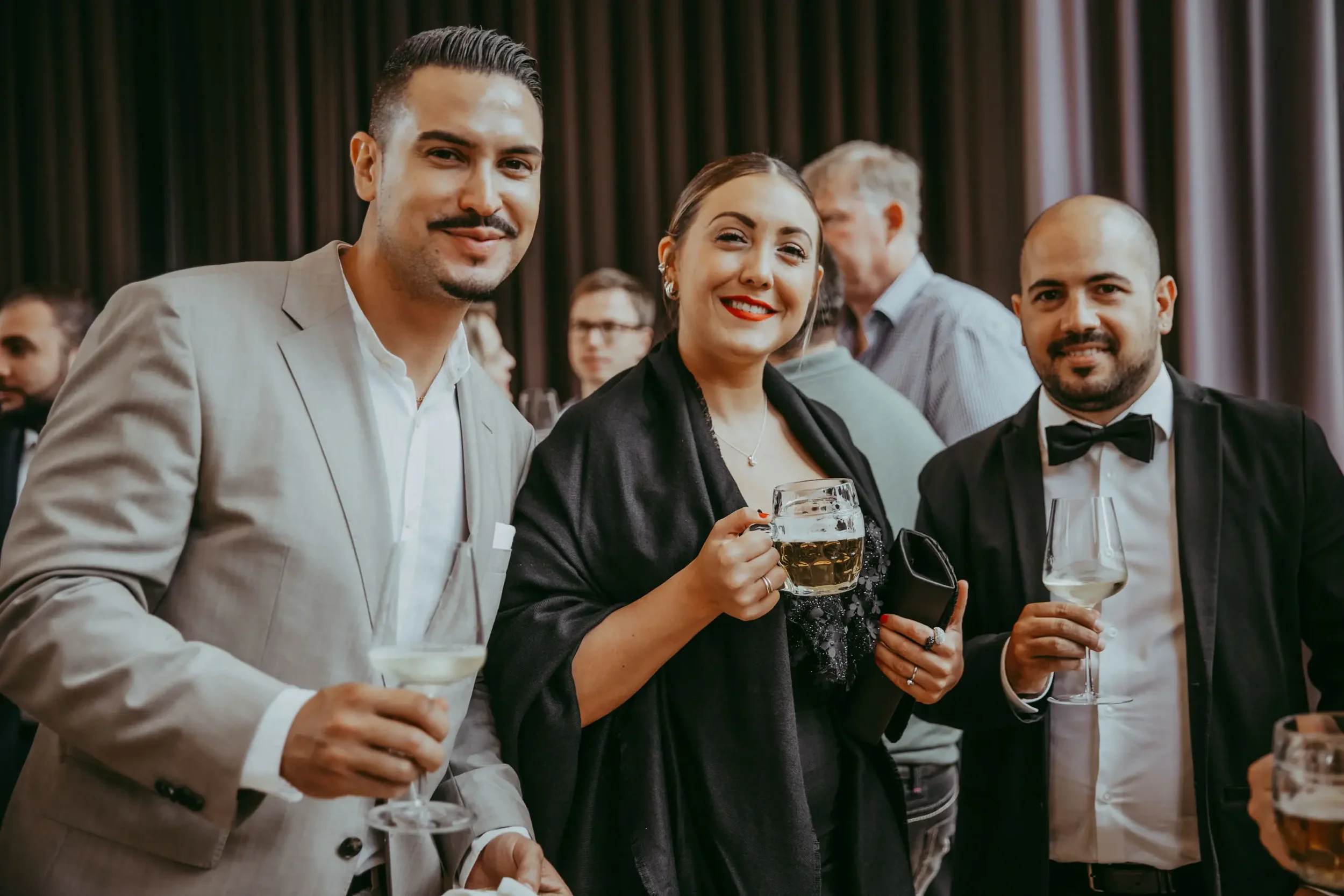 Menschen auf einer Feier mit Getränken, eine Frau lächelt in die Kamera, während sie ein Glas Bier hält, im Hintergrund sind weitere Gäste, die bei einer Feier sind.