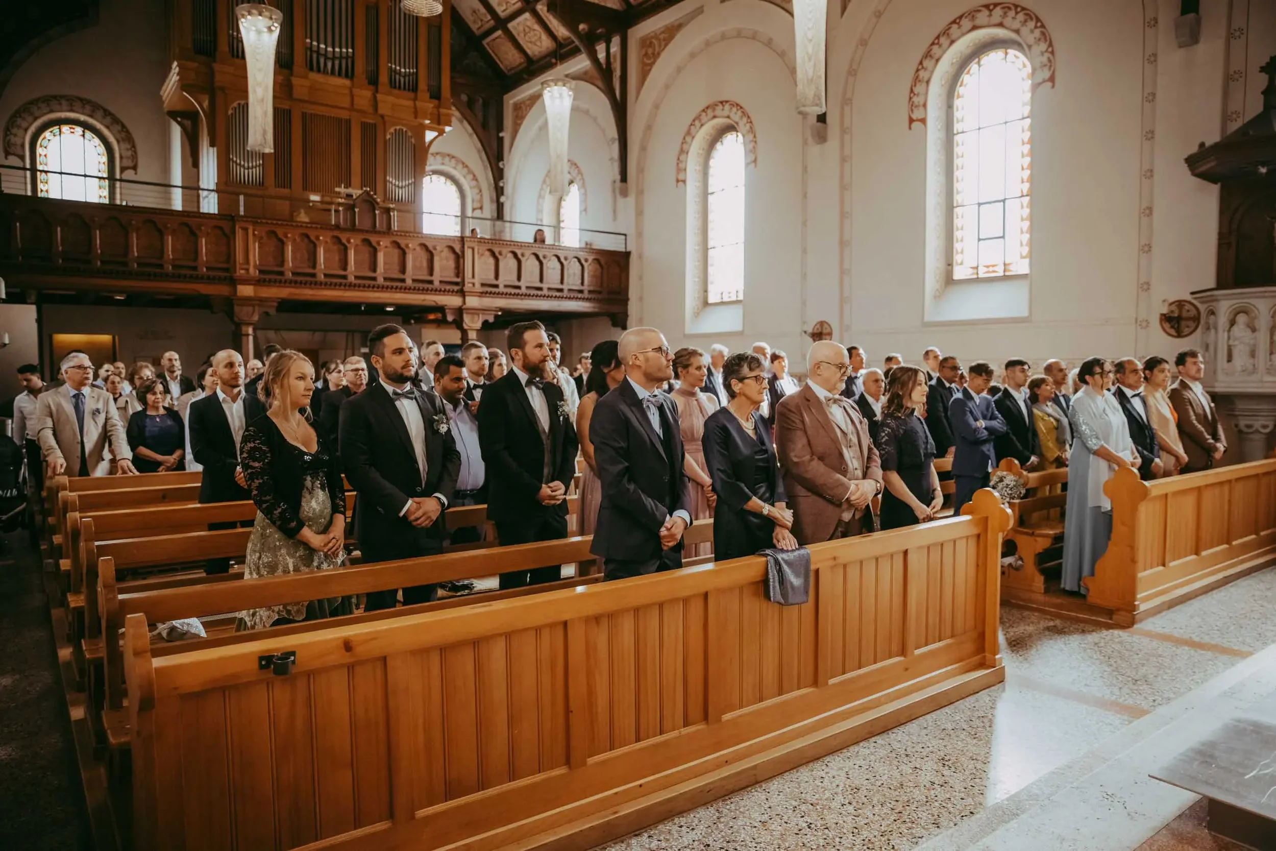 Menschen in festlicher Kleidung sitzen und stehen in einer Kirche während einer Zeremonie, mit hohen Fenstern und christlichen Dekorationen im Hintergrund.