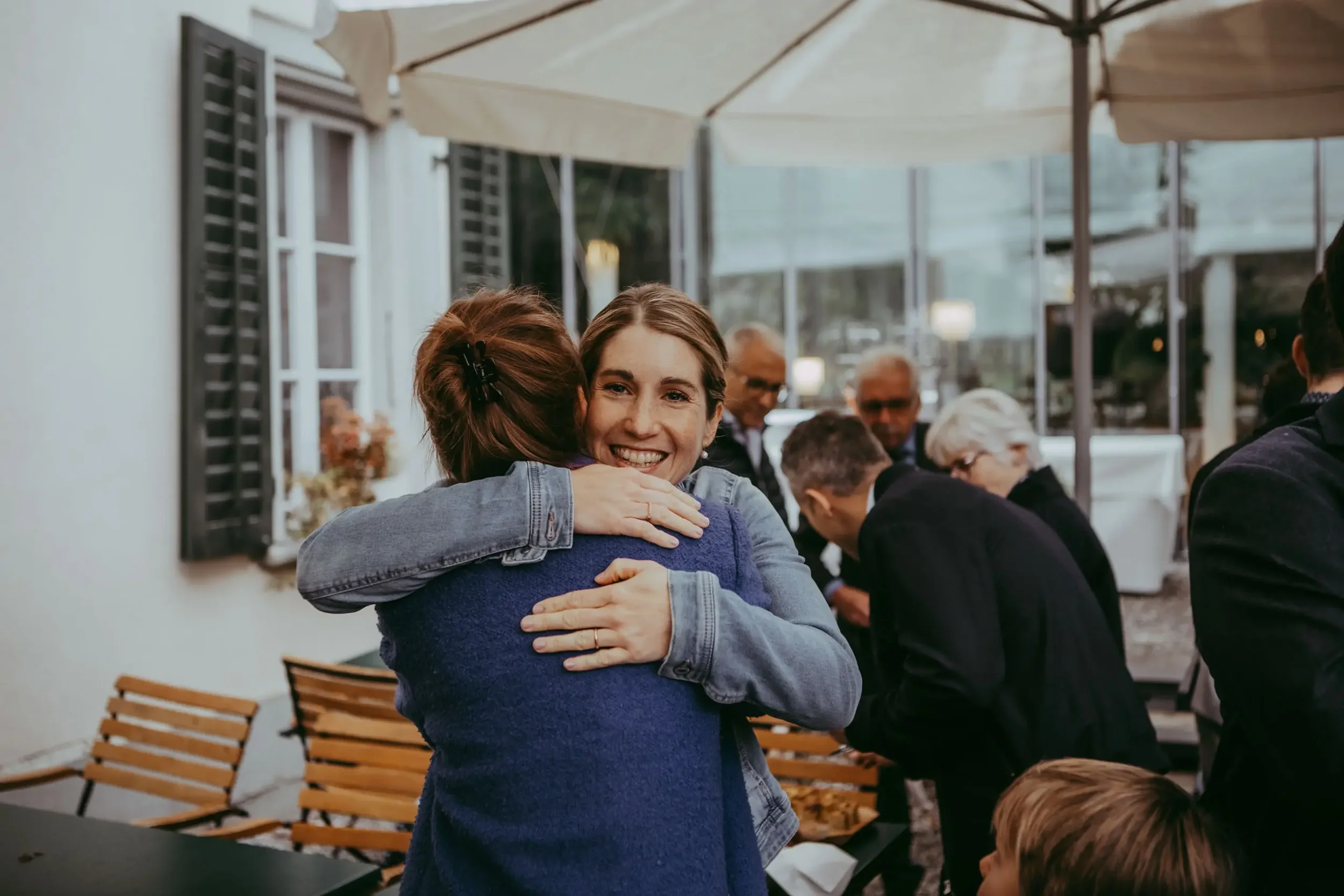 Zwei Frauen umarmen sich freudig in einem Cafegarten, während im Hintergrund mehrere ältere Menschen an einem Tisch sitzen.