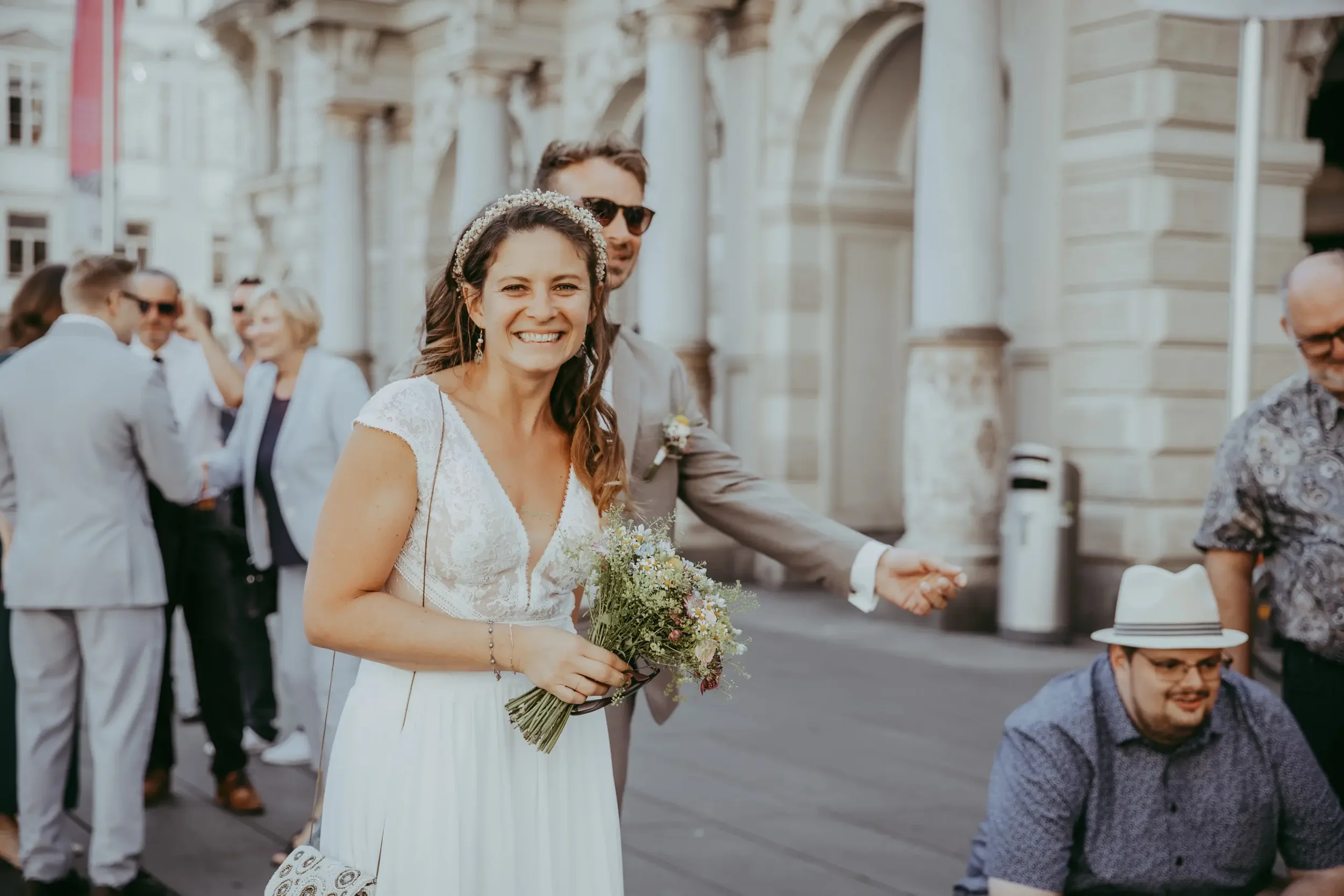 Eine lachende Braut in weißem Kleid hält einen Blumenstrauß und geht auf einer Hochzeit. Hinter ihr stehen Gäste in formeller Kleidung, die an einem eleganten Gebäudeteil in der Stadt feiern.