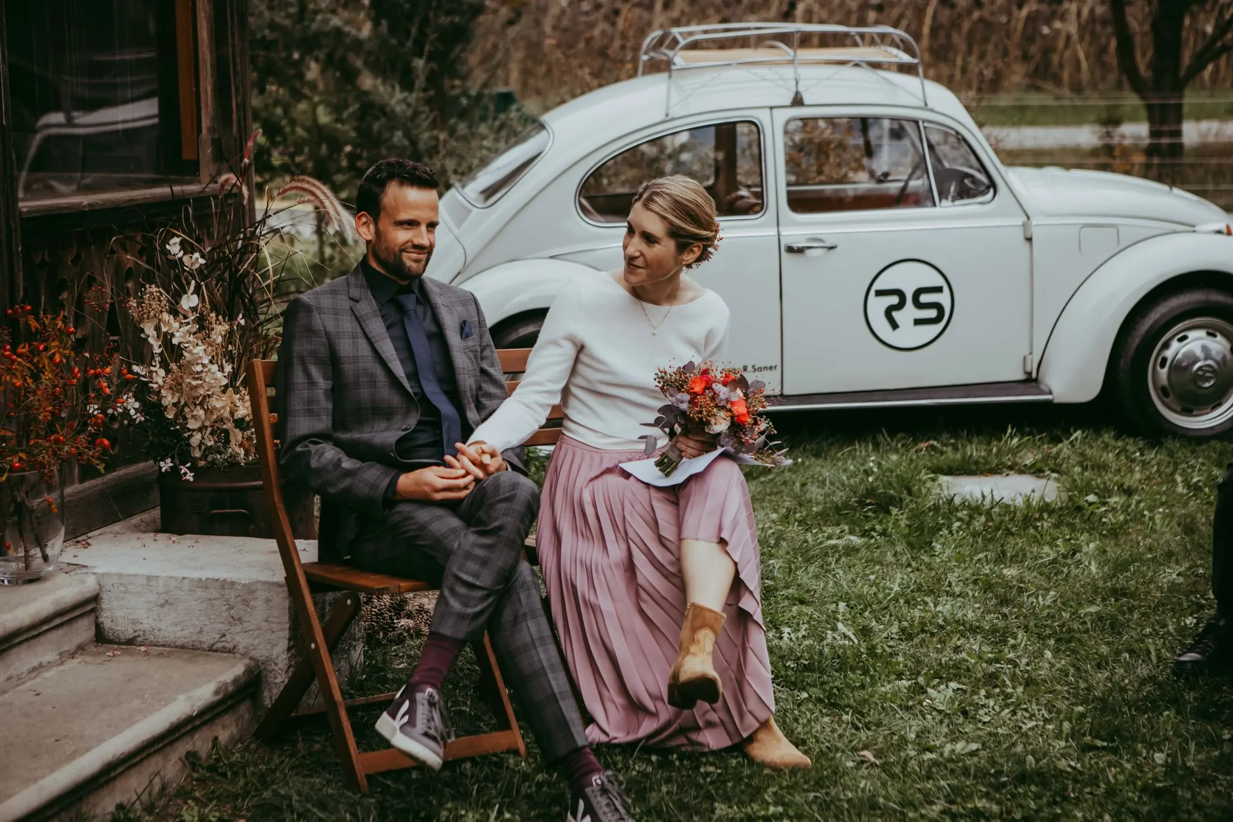 Ein Mann und eine Frau sitzen auf einer Bank im Garten, sie halten Händchen und schauen sich an, im Hintergrund ist ein alter weiße VW Käfer mit einem Dachgepäckträger zu sehen.