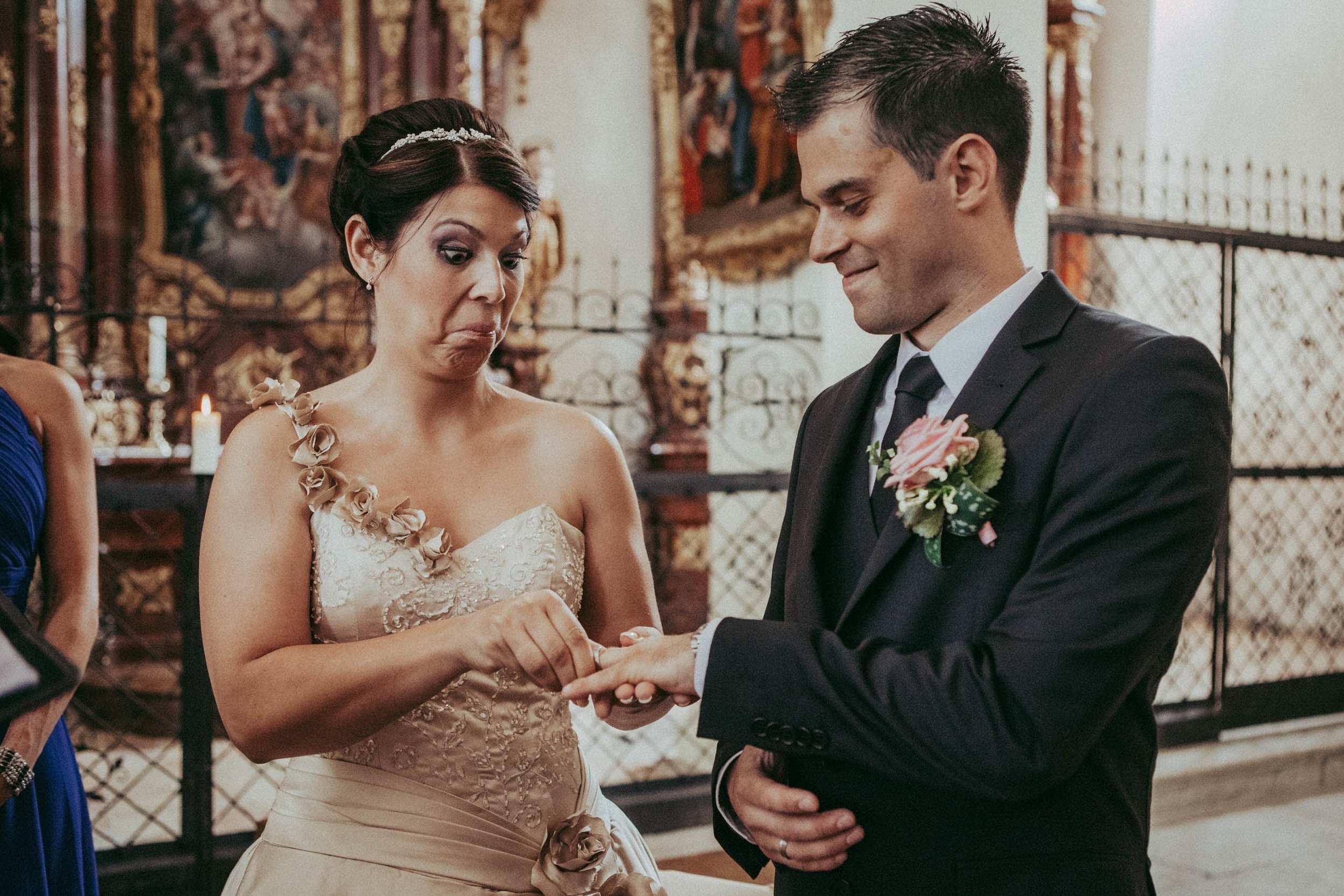 Ein Brautpaar bei einer Hochzeit, die Hand der Braut wird vom Bräutigam gehalten, im Hintergrund kunstvolle Wanddekorationen.