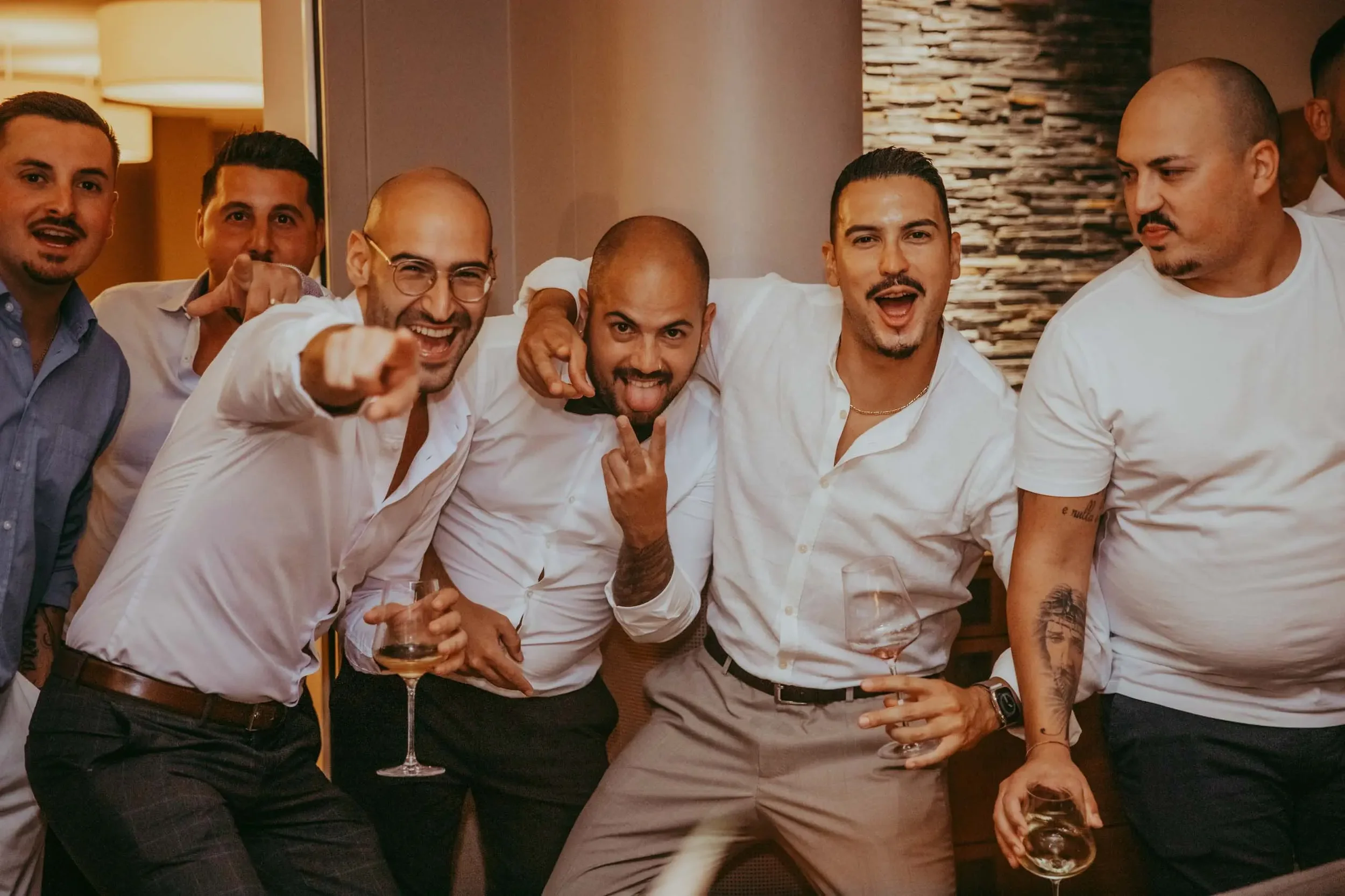 Eine Gruppe von sieben Männern, die bei einer Party oder Zusammenkunft zusammenstehen. Sie lachen, halten Getränke in der Hand und zeigen gewisse Gesten. Die Stimmung ist fröhlich und gesellig.