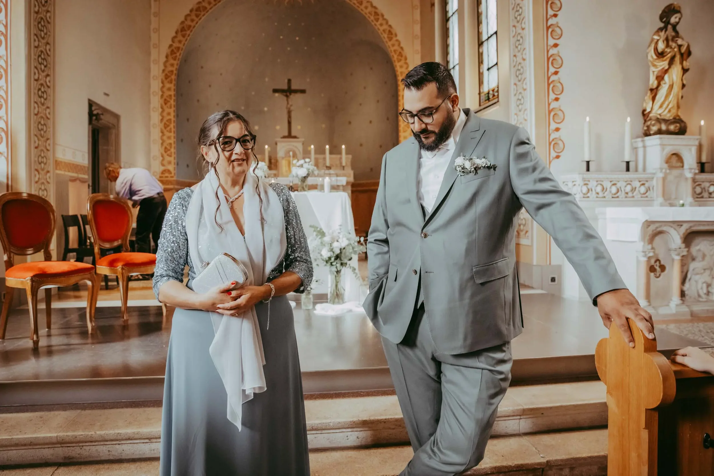 Zwei Menschen in formeller Kleidung, eine Frau und ein Mann, stehen in einer Kirche vor dem Altar. Der Hintergrund zeigt einen Kreuz in der Mitte, Kerzen und religiöse Statuen.