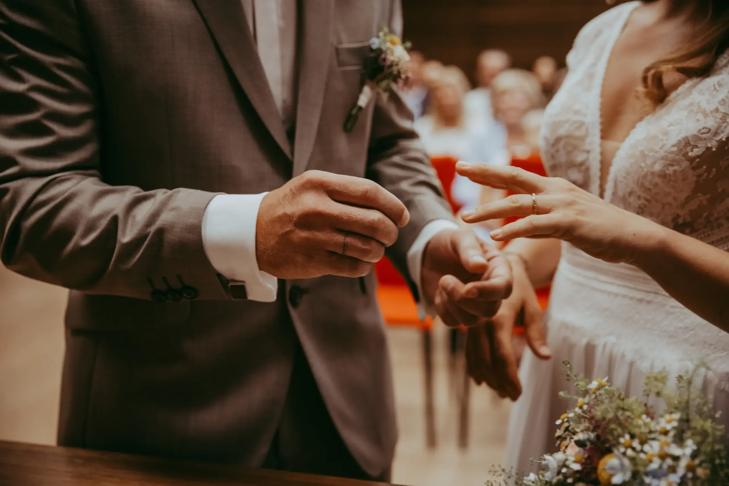 Ein Brautpaar tauscht sich bei ihrer Hochzeit die Ringe aus, mit Gästen im Hintergrund.