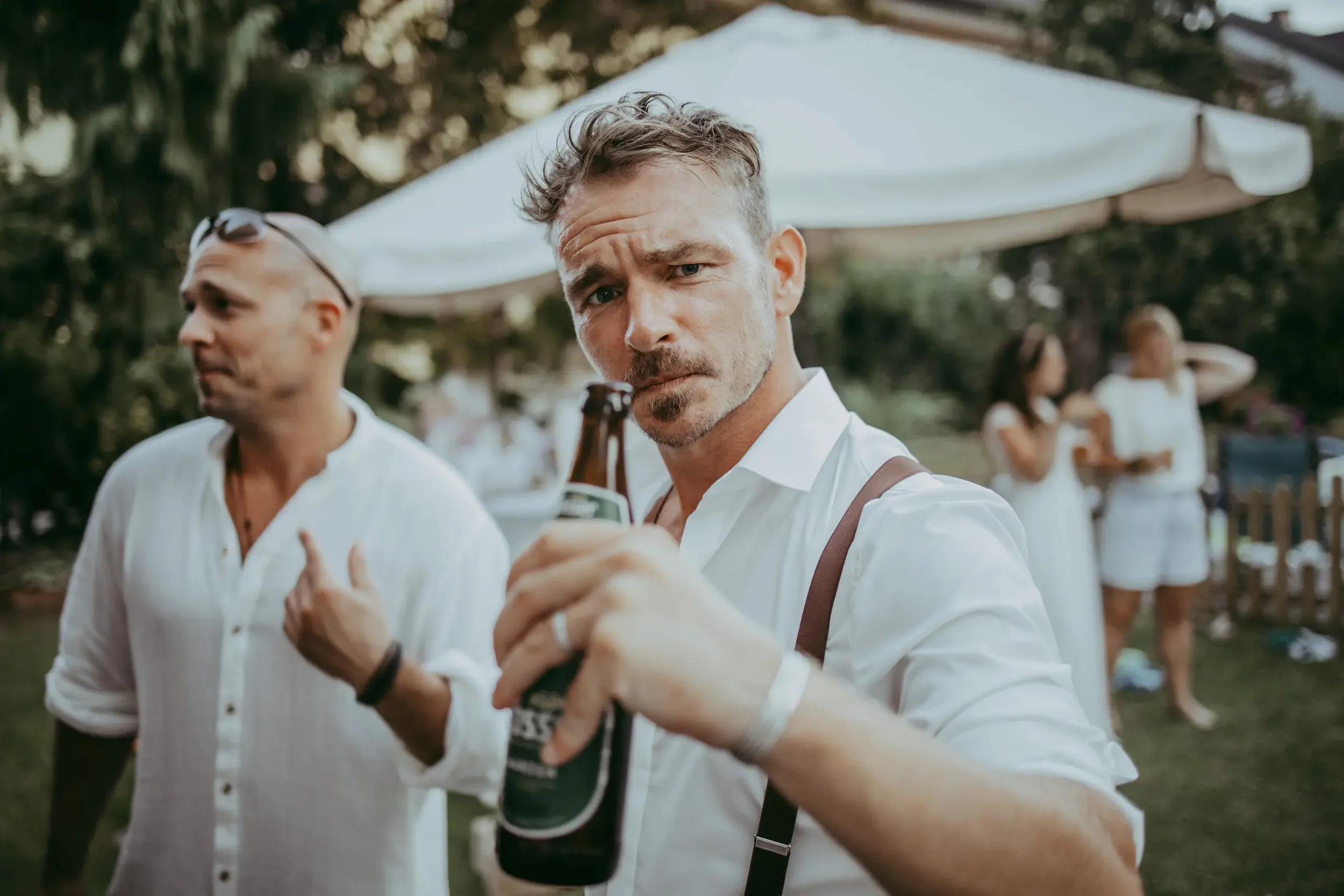 Junger Mann mit braunen Haaren und Bart, der ein Bierglas in der Hand hält, bei einer outdoor Party. Im Hintergrund weitere Menschen und eine weiße Sonnenschirm. Es sieht nach einem sonnigen Tag aus.