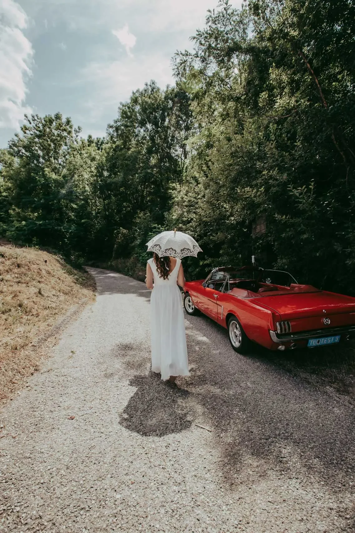 Eine Frau in einem weißen Kleid läuft auf einem ländlichen Weg, hält einen weißen Schirm und steht neben einem roten Oldtimer. Die Umgebung ist grün mit Bäumen und einer Wolke am Himmel.