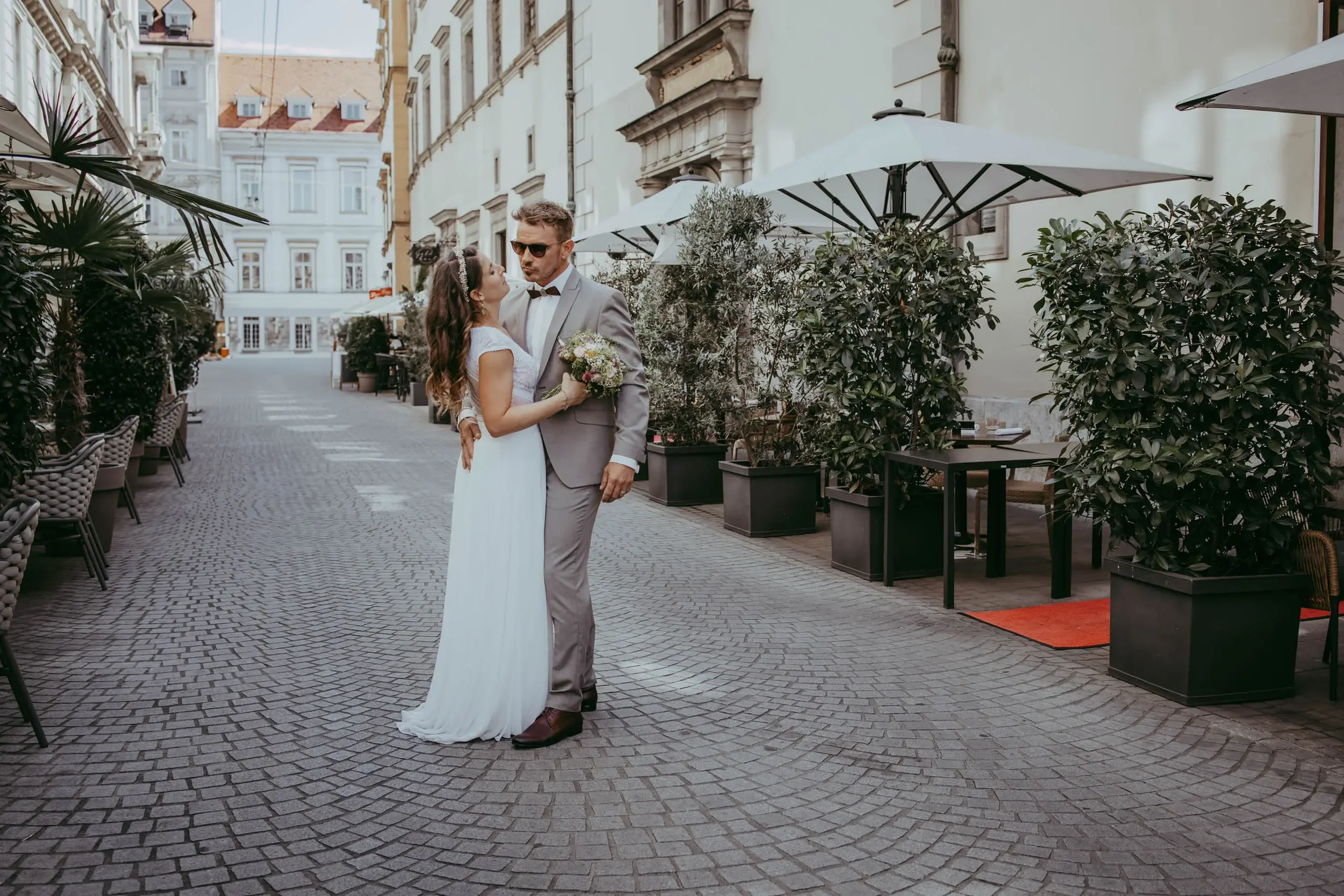 Ein Brautpaar in formeller Kleidung auf einer gepflasterten Straße in einer europäischen Stadt, umgeben von Gebäuden und Büschen, beim Fotografieren.