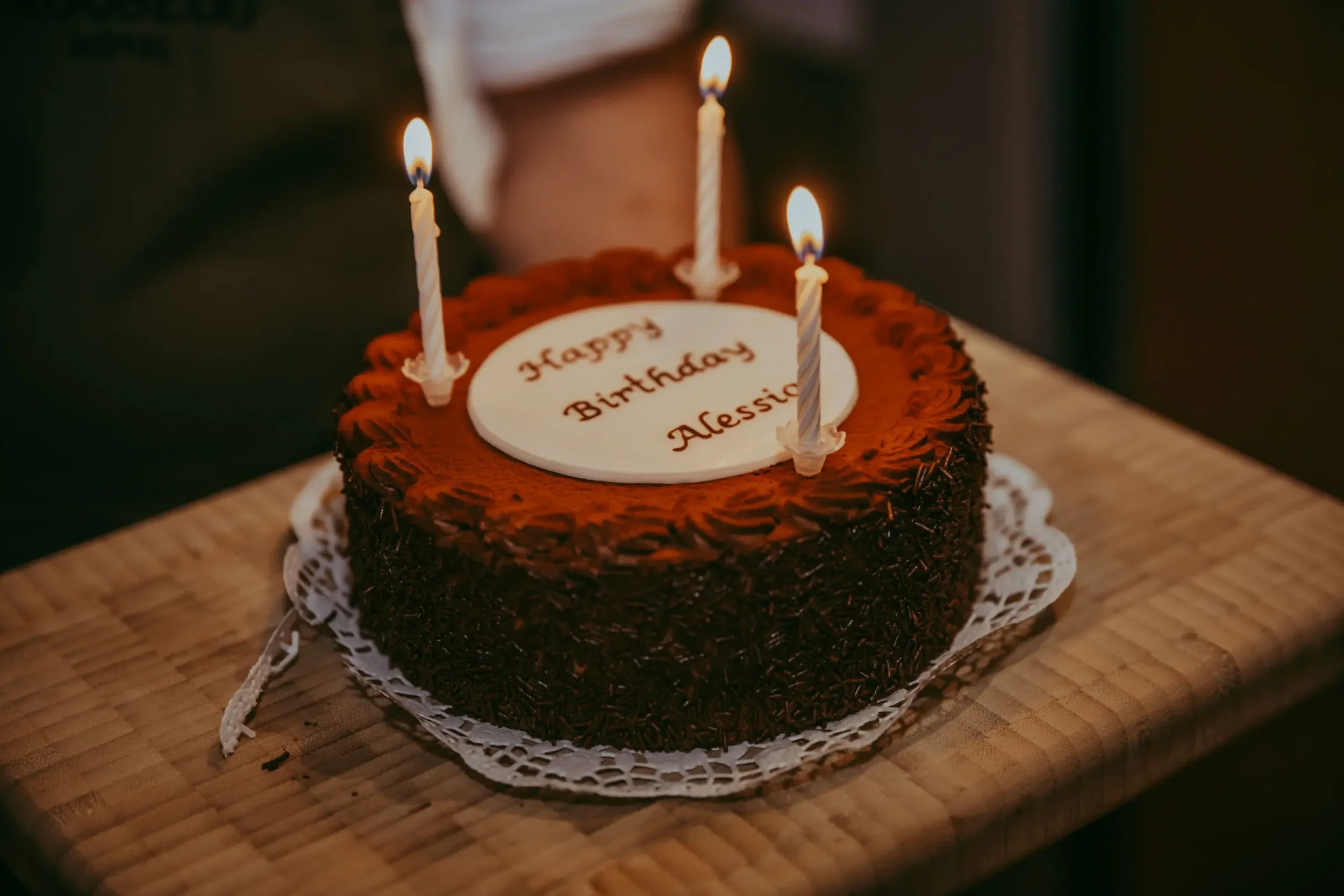 Ein runder Schokoladenkuchen mit drei brennenden Kerzen und einer weißen Zuckerglasur mit der Aufschrift 'Happy Birthday Alessia' auf einem Holztablett.