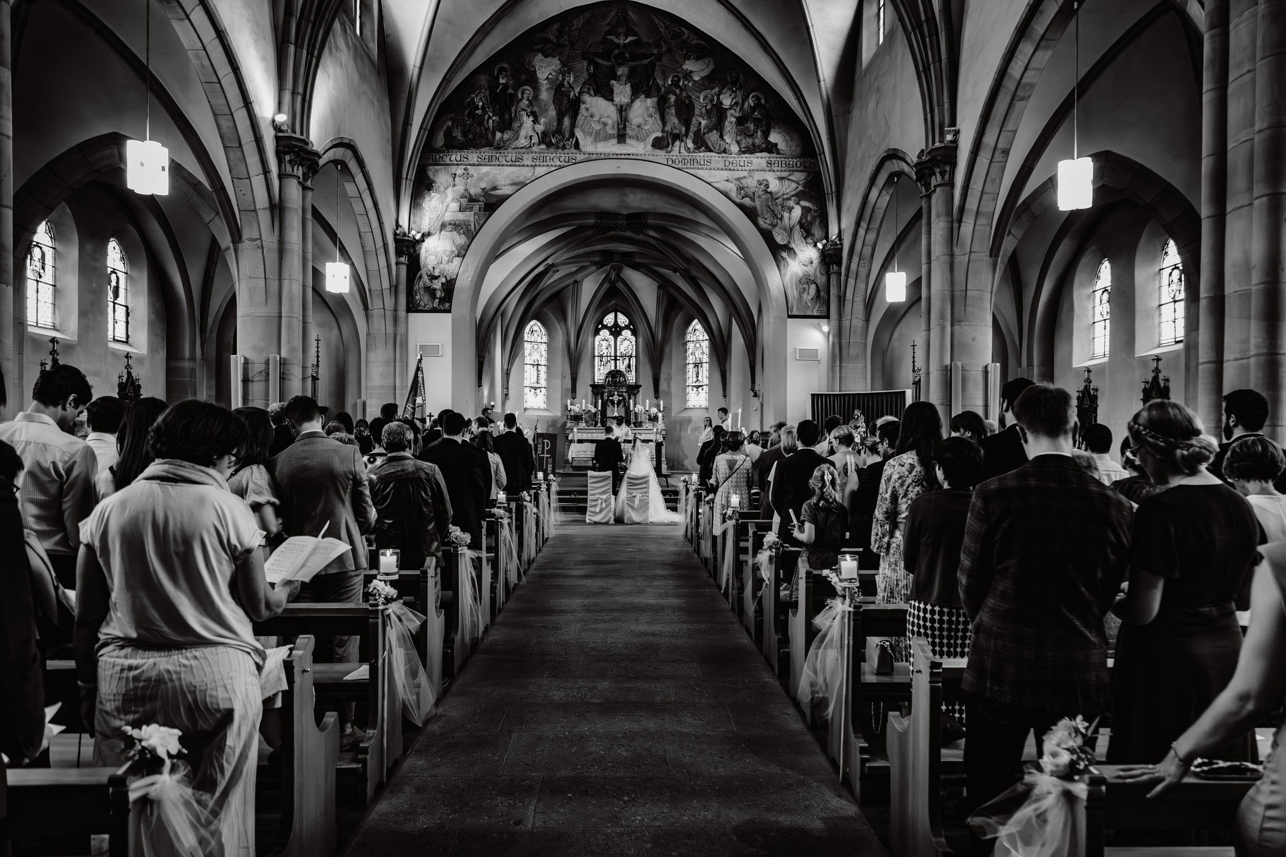 Schwarz-Weiß-Foto einer Kirche während einer Hochzeitszeremonie, mit vielen Gästen, die vor dem Altar sitzen, und einem Brautpaar im Mittelpunkt.