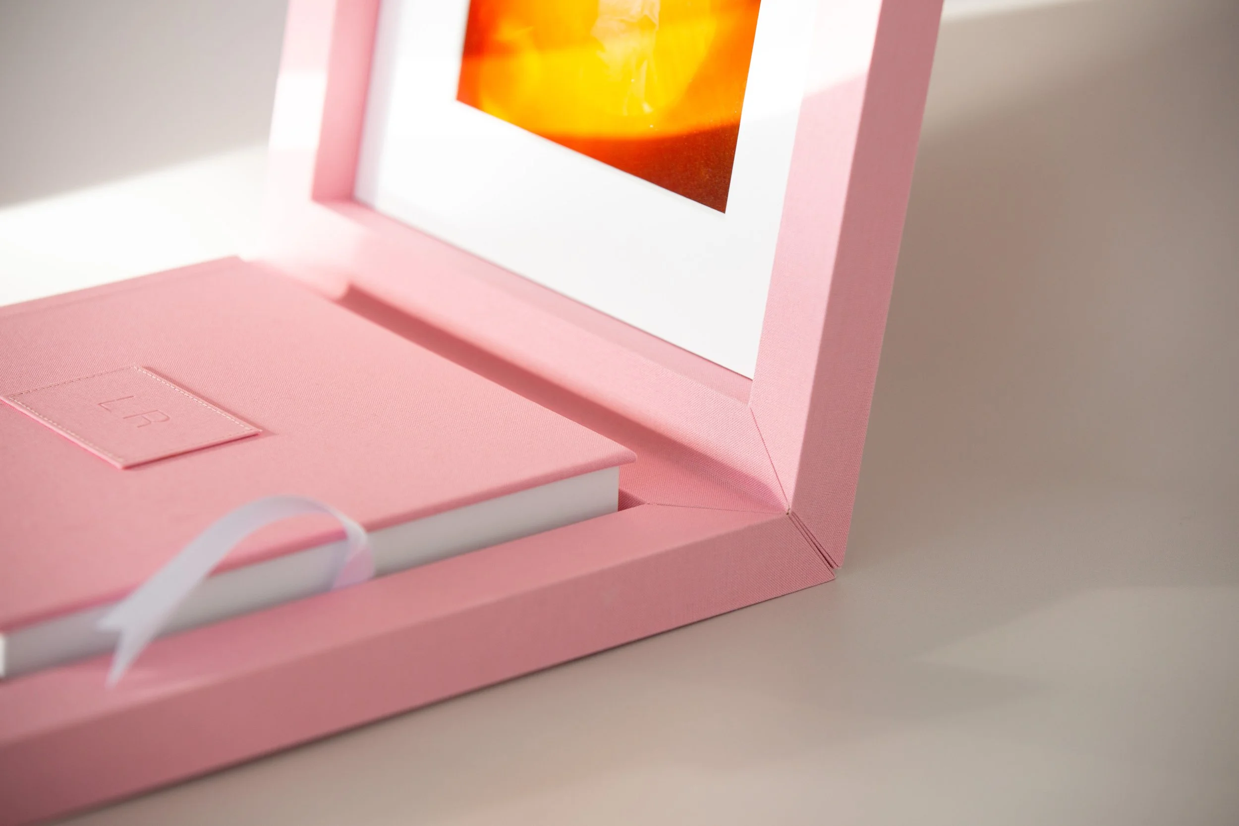 Ein rosa Geschenkbox mit einem geöffneten Deckel, in dem sich ein gerahmtes Bild mit orange-weißem Muster befindet, auf einem weißen Hintergrund.