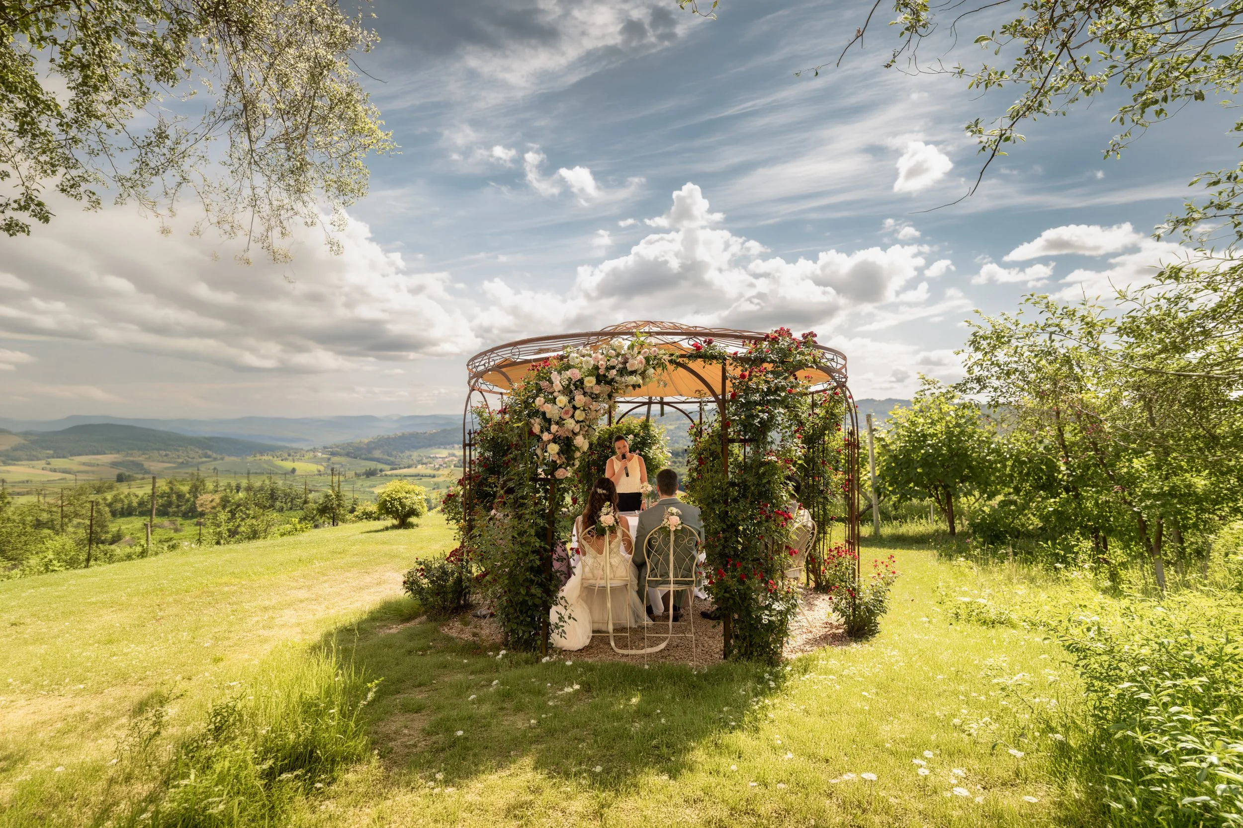 Hochzeit im Grünen mit einer Zeremonie unter einer Blumenüberdachung im Freien auf einer Wiese mit Blick auf die Landschaft und den Himmel