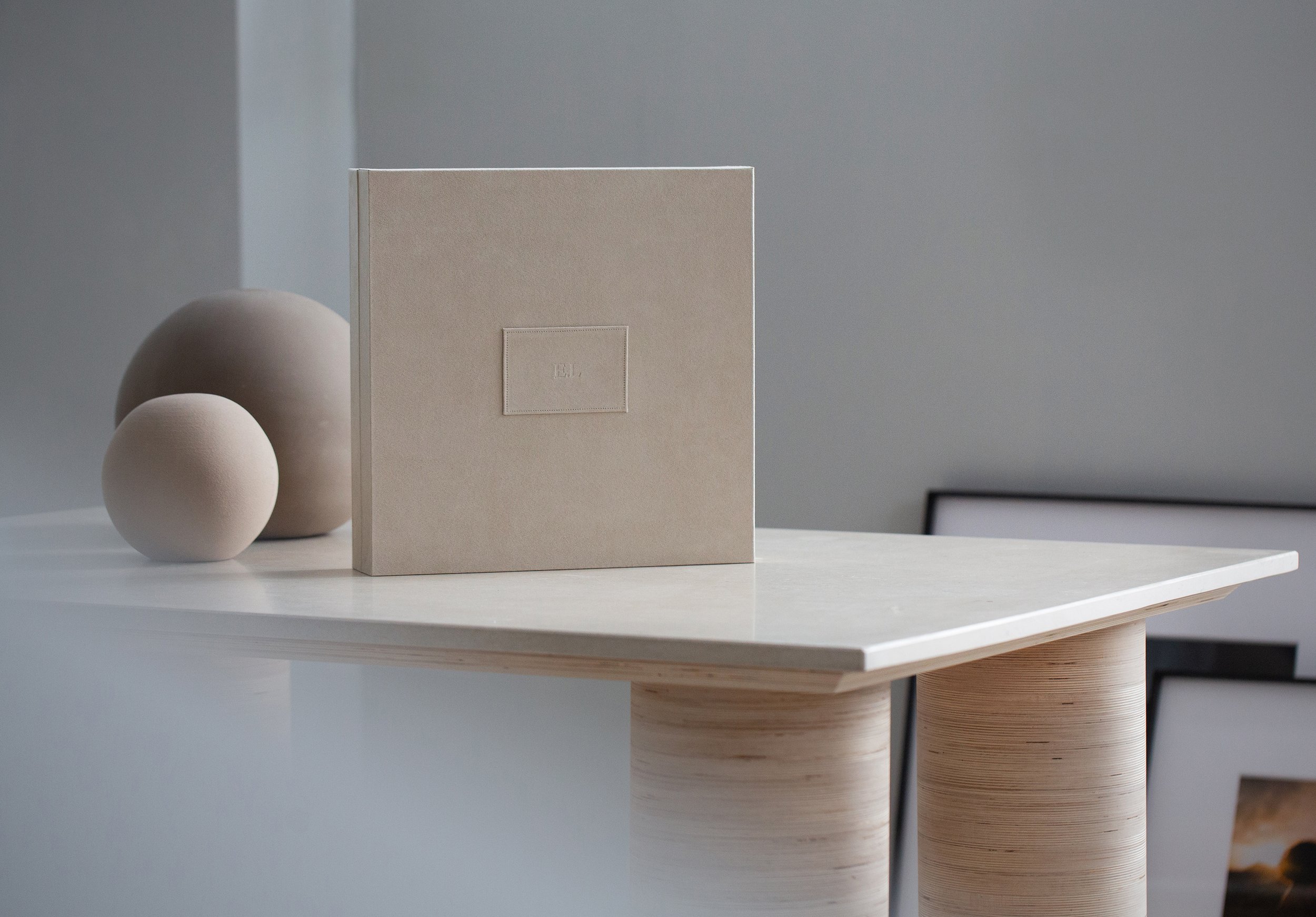 Eine weiße oberfläche mit zwei weißen, runden Keramikkugeln auf der linken Seite und einem beige Buch mit Monogramm "E.L." auf der Mitte. Im Hintergrund eine graue Wand.