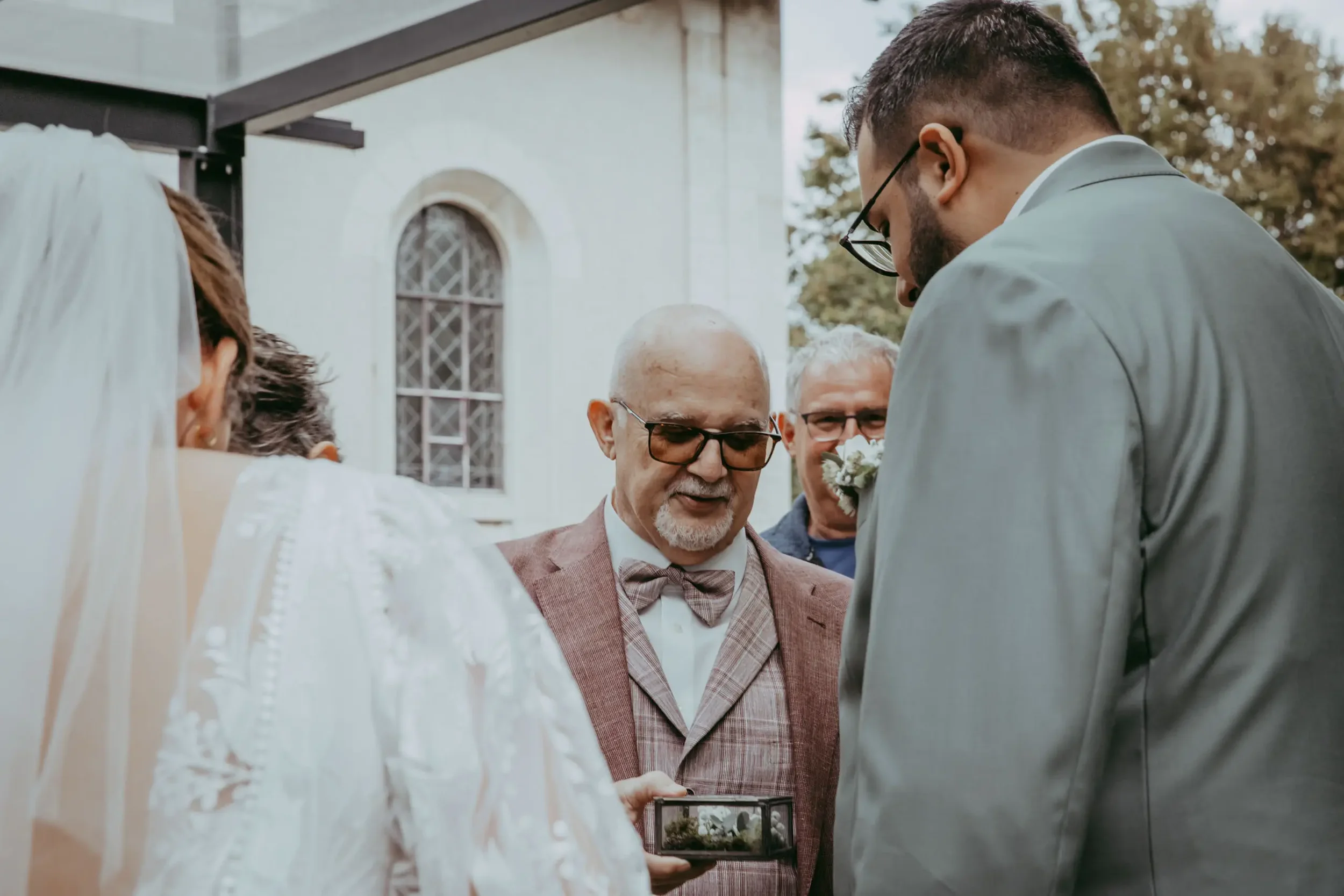 Auf diesem Bild sehen wir eine Hochzeitszeremonie vor einer Kirche im Freien. Mehrere Personen, darunter ein älterer Mann mit Brille, ein jüngerer Mann mit Brille und ein Mann mit dunklem Haar, sind beteiligt. Ein Mann in einem Anzug hält ein kleines