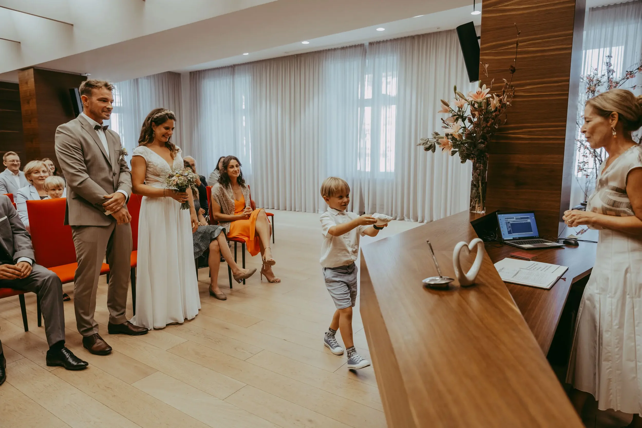 Hochzeit im Festsaal, Braut und Bräutigam stehen vor der Trauzeugen, während Gäste sitzen und zuschauen, eine Frau steht am Empfangstisch.